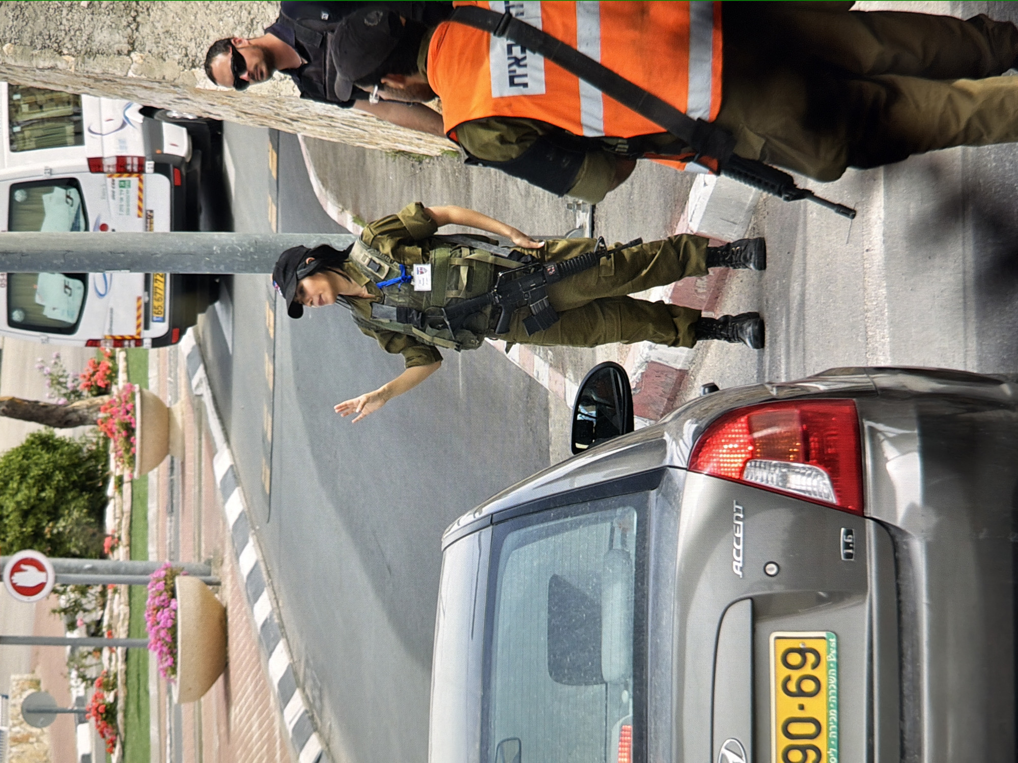 The image depicts an outdoor scene with several individuals, a car, and a building facade. In the foreground, a gray Hyundai Accent car is parked. Behind the car, three people are visible. The person on the left is a woman in military uniform, wearing a cap and a tactical vest, holding a rifle. She is looking to her right. To her right, a man is wearing a dark shirt and sunglasses. Partially visible in the right foreground is another person wearing an orange vest over a uniform, with a rifle slung across their chest. The background shows a street with a white van, a sidewalk with planters containing pink flowers, and a stone wall. The presence of Hebrew writing on the car's license plate and on the orange vest suggests the location is likely in Israel, not Kyiv, Ukraine.