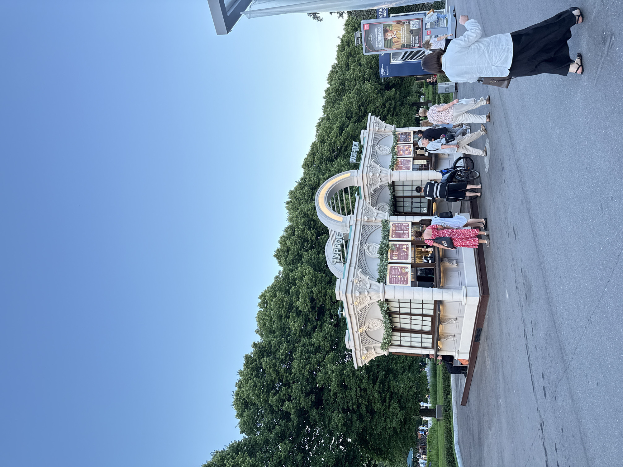 The image depicts an outdoor scene in Moscow, Russia, likely in a park or public square, with a clear blue sky overhead. In the foreground, people are walking on a paved area. A white kiosk with ornate architectural details, including columns and decorative trim, is the central subject. The kiosk appears to be a food or beverage stand, with menus displayed on its windows and a few customers and staff interacting around it. Behind the kiosk, a dense line of mature green trees forms a backdrop. To the right of the kiosk, a large digital billboard displays an advertisement. The overall atmosphere suggests a pleasant day in a public gathering space.