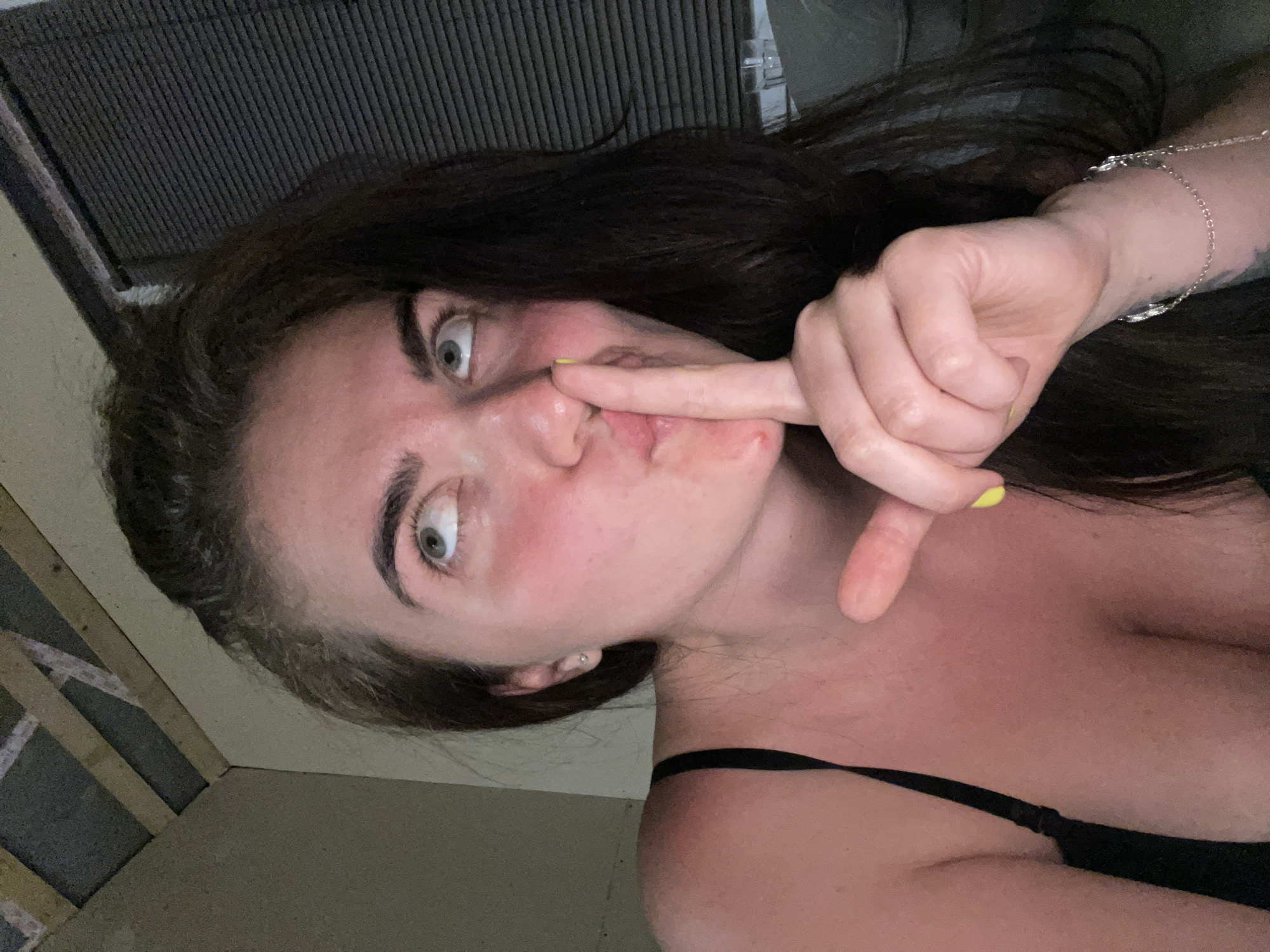 A medium close-up shot captures a woman with dark brown hair and fair skin, her face tilted upwards and to the left. She is wearing a black bra and has a light pink flush across her cheeks and chest, indicative of a sunburn. Her right index finger is raised to her lips in a shushing gesture, and her eyes are wide open, looking upwards. Her fingernails are painted yellow. In the background, the ceiling shows exposed wooden beams and what appears to be insulation. To the right, a window with closed blinds is visible, and a small glass is placed on a surface. A silver bracelet adorns her wrist, and a tattoo is partially visible on her forearm. The overall lighting suggests an indoor setting, possibly at night or in a dimly lit room. The location is not determinable from the image.