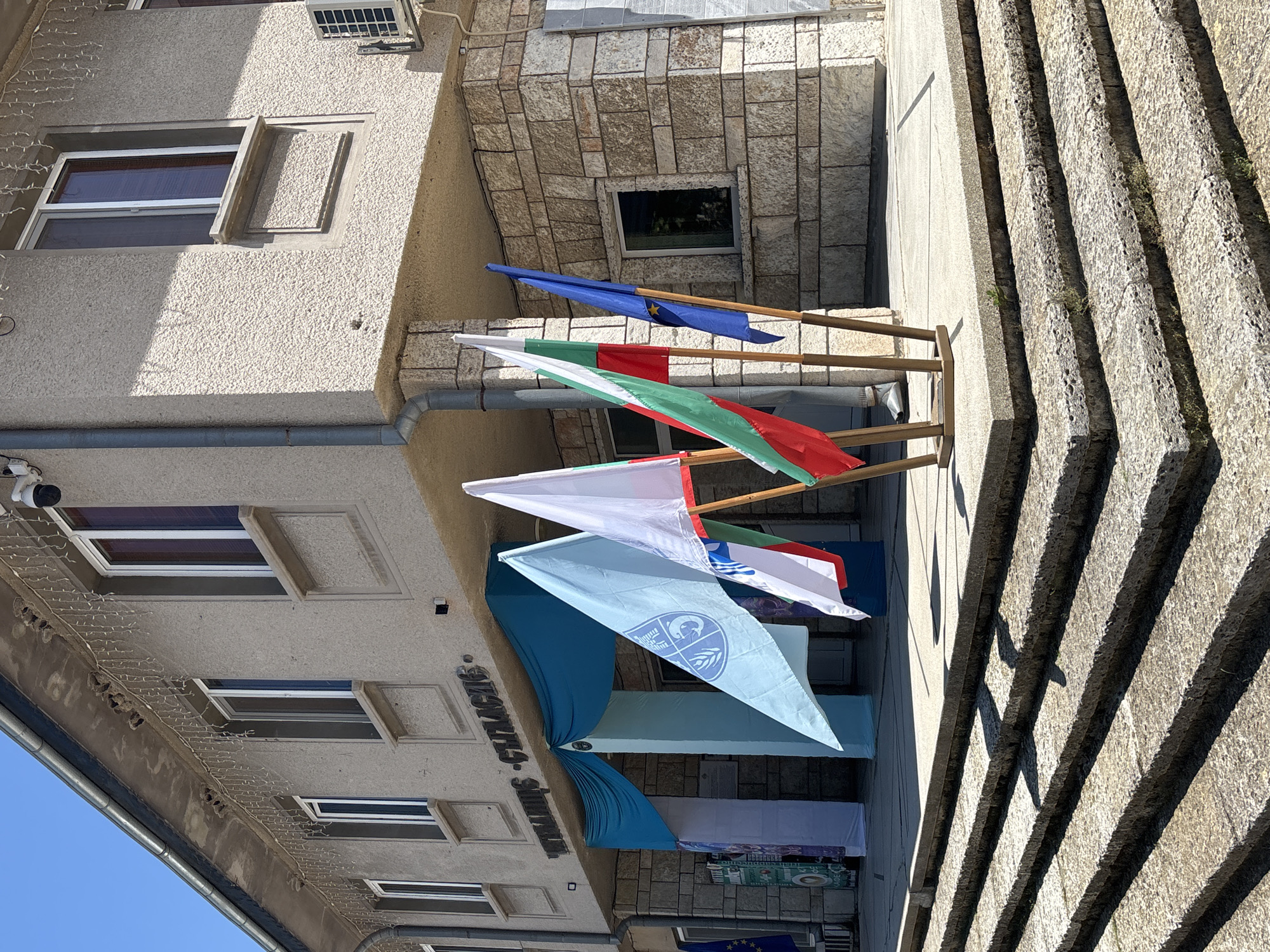 The image depicts the entrance to a building in Kavarna, Bulgaria, likely a municipal or cultural center, as indicated by the Cyrillic inscription above the entrance which reads "ЧИТАЛИЩЕ - СЪГЛАСИЕ" (Chitalishte - Saglasie, meaning "Reading Room - Harmony"). The building has a light-colored stucco facade with stone accents around the lower level and entrance. Several flags are displayed prominently on wooden stands in front of the building. These include the Bulgarian flag (white, green, red), the European Union flag (blue with yellow stars), and another flag with a light blue background and a circular emblem featuring stylized figures and waves. The entrance is adorned with blue drapery. Steps made of rough-hewn stone lead up to the building's entrance. The sky is clear and blue, suggesting a sunny day. The overall scene is festive, possibly in anticipation of or during a public event.