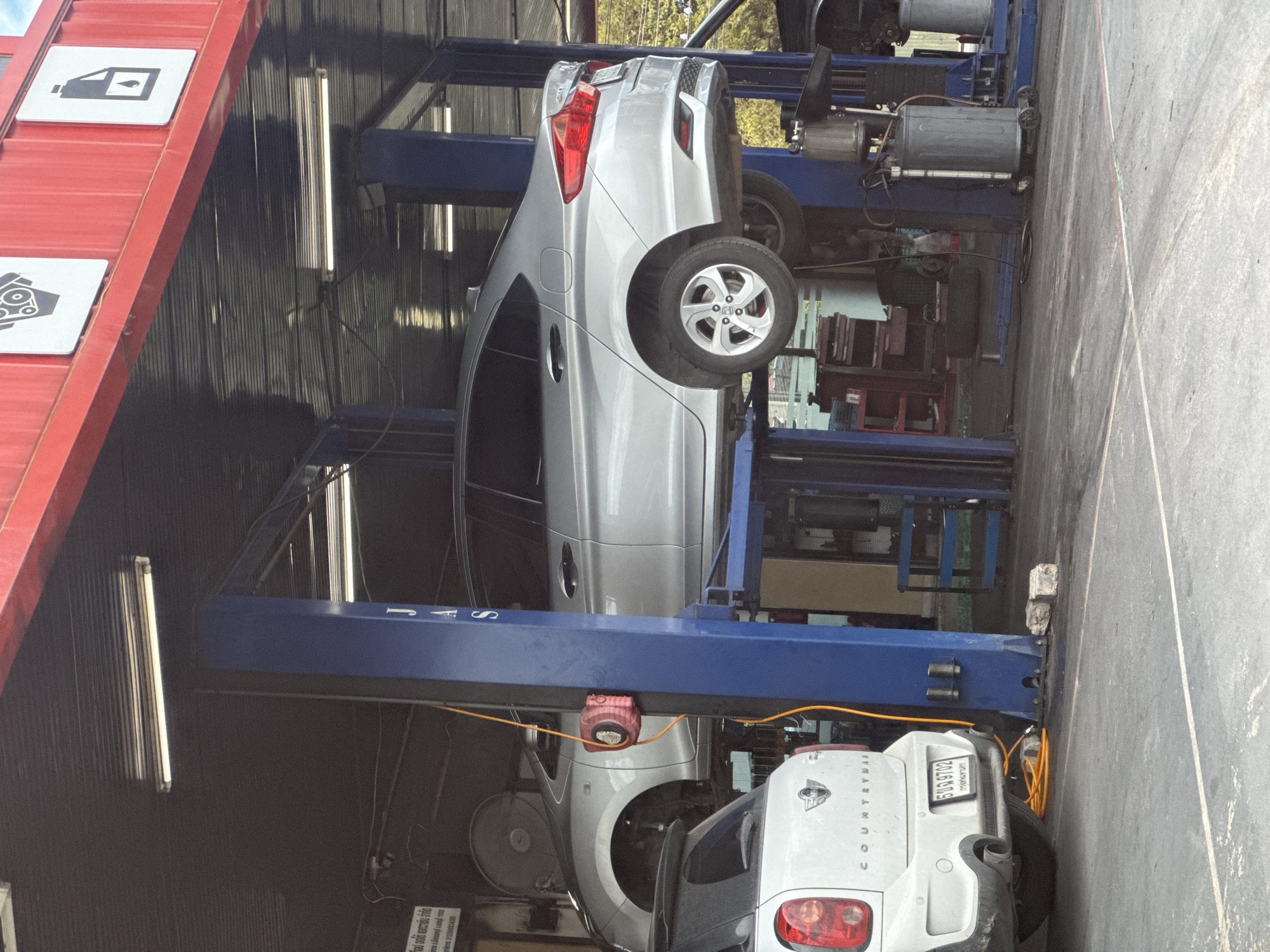 The image depicts a car repair shop in Nonthaburi City Municipality, Thailand. Two cars are visible: a white Mini Cooper Countryman is parked on the ground in the foreground, and a silver sedan is elevated on a hydraulic lift in the background. The repair shop features blue hydraulic lift pillars, fluorescent lighting, and red corrugated metal siding on the exterior. Various tools and equipment, including a red coiled hose and a gray oil drain can, are present in the scene. The ground is paved with asphalt. The overall environment suggests an active automotive service facility.