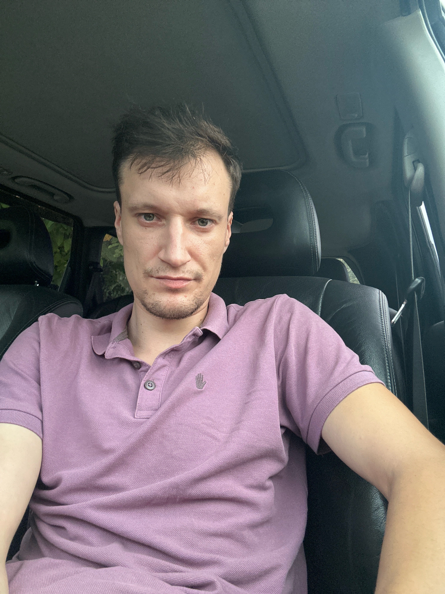 A man is sitting in the driver's seat of a car, looking at the camera. He has short, brown hair and is wearing a purple polo shirt with a small hand logo on the left chest. The interior of the car is visible, with black leather seats and a sunroof. The background outside the car appears to be foliage, suggesting an outdoor setting, possibly in Shatava, Ukraine.