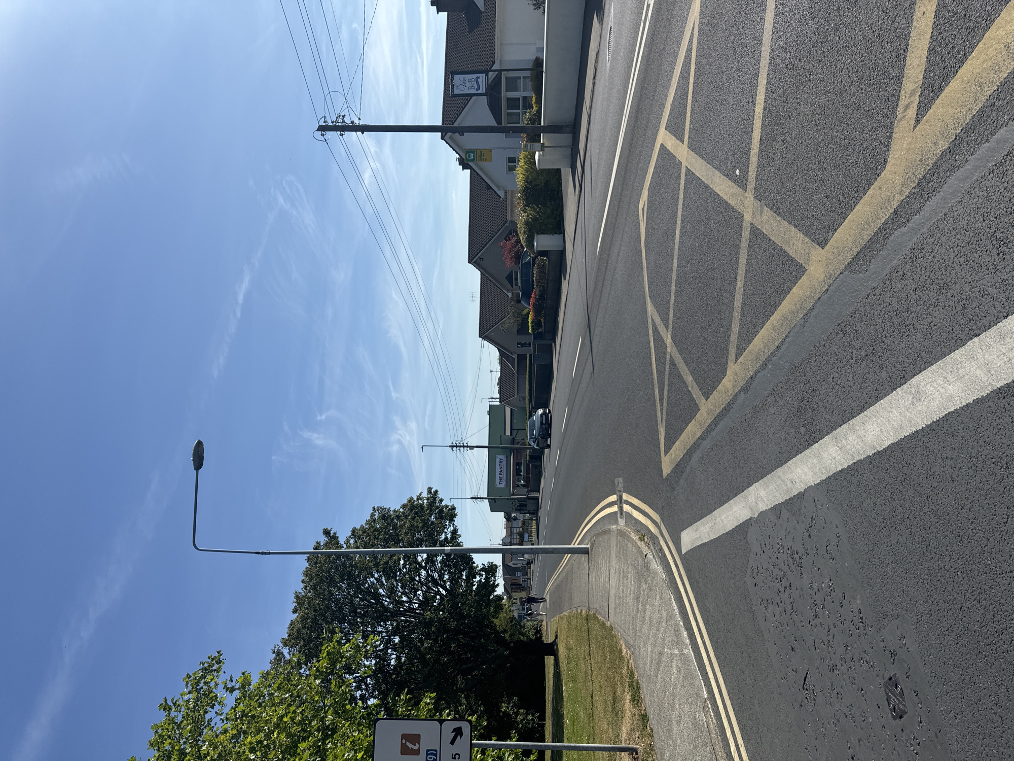 The image shows a street scene on a sunny day in the city of Waterford, Ireland. The foreground features a paved road with double yellow lines and a white line indicating a lane divider. A yellow box junction is visible on the right side of the road. To the left, a concrete sidewalk runs alongside a grassy area with a large tree. A signpost with a blue directional sign containing numbers and arrows stands on the left.
In the mid-ground, the road continues with a few cars driving. Buildings line the right side of the street, including what appears to be a business labeled "THE PANTRY" with a green facade and a sign for "Vee B&B" above a white house. Power lines are visible overhead, supported by a utility pole. The sky is blue with some wispy white clouds. Two people are walking on the sidewalk in the distance. The overall impression is a typical suburban or semi-rural street in Ireland.