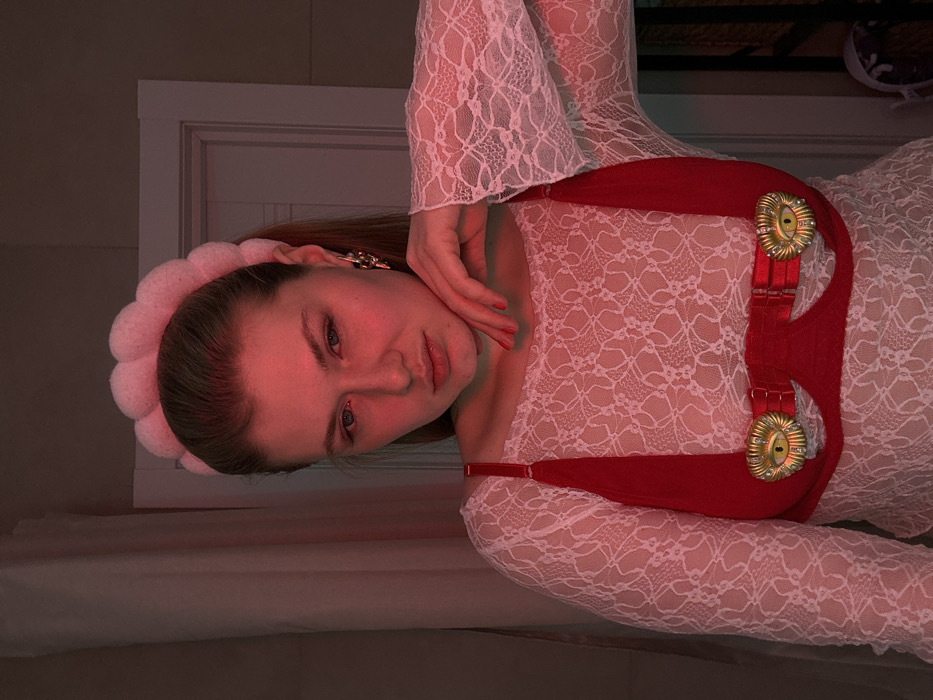A young woman lies on a surface, angled to the left. She wears a white lace top with flared sleeves, a plush pink headband, and ornate gold earrings. A red, structured harness with decorative gold buckles adorns her chest. Her left hand gently touches her face, drawing attention to her lips and eyes. The scene is bathed in a warm, reddish light. A white wall and part of a window frame are visible in the background.