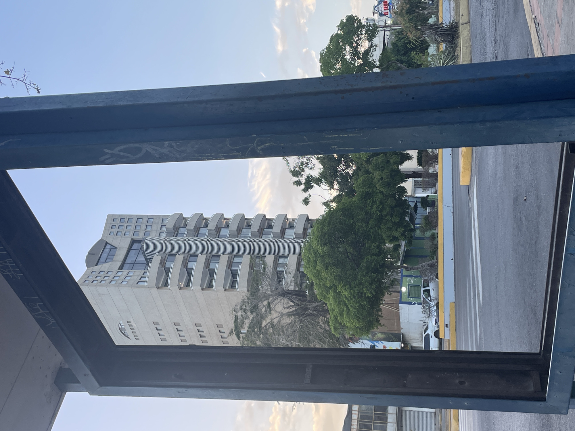 The image, taken in Porlamar, Venezuela, shows a high-rise building through a metal structure, possibly a bus stop or similar shelter. The building is light beige, with a modern design featuring multiple balconies and a glass section towards the top. Trees are visible on the right side of the building, partially obscuring the view. A road with white lane markings and yellow curb is seen to the right of the trees. Cars are parked alongside the road. The sky is light blue with scattered clouds. Graffiti marks part of the metal structure framing the view. Lower buildings appear in the bottom.