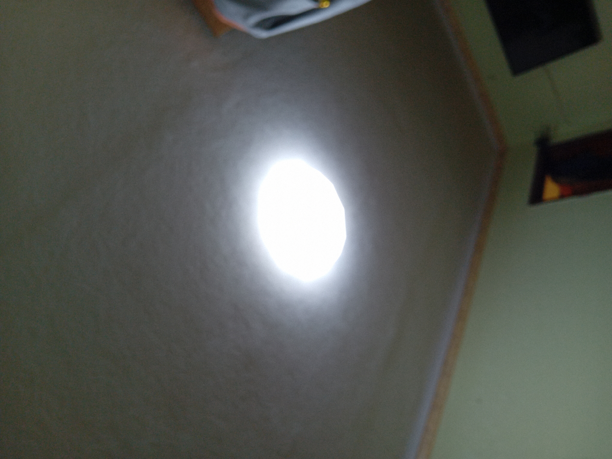 The image shows the ceiling of a room with a bright, circular light fixture in the center. The ceiling appears to be textured and a light gray color. The walls of the room are a pale green. There is a wooden trim or molding where the ceiling meets the walls. In the upper right corner of the image, part of a gray garment is visible, possibly clothing hanging or on a person. The bottom portion of the image shows a dark rectangular object, likely a television, mounted on the wall. To the left of the television, a doorway is visible, with a hint of yellow light emanating from within. The image is taken from a low angle, looking upwards. No specific details about the city of Chuhuiv, Ukraine, are discernible from the image itself.