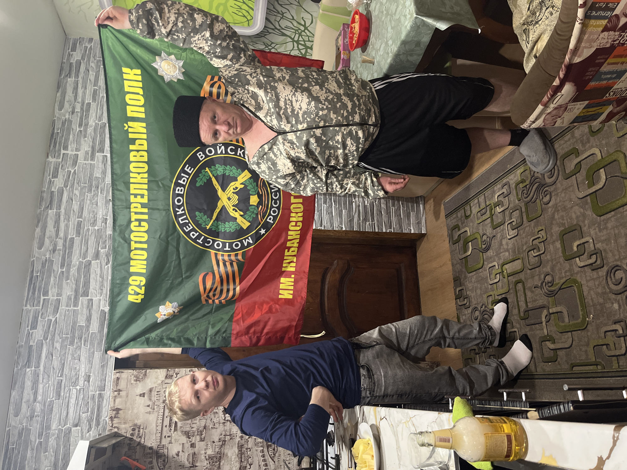 Two men are holding a flag. The flag is colored green, red, and black. It depicts crossed rifles, stars, and the words "429 МОТОСТРЕЛКОВЫЙ ПОЛК". The man on the left has blonde hair, is wearing a dark blue shirt, and gray jeans. The man on the right is wearing a camouflage jacket, black shorts, and a black hat. A table with food is visible near the men. The location appears to be indoors, likely a home in Vesyoloye, Russia, based on the flag's markings.