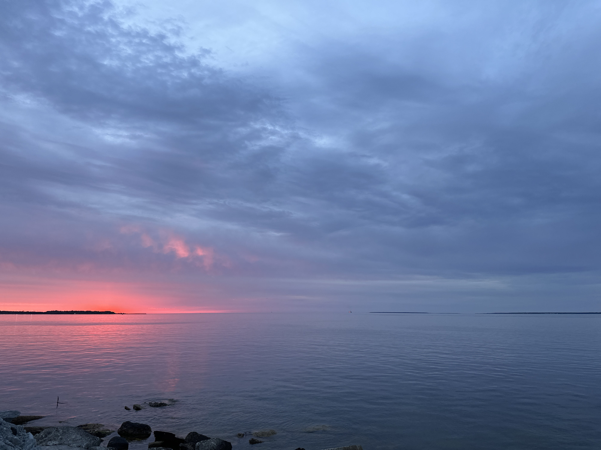 The image depicts a serene seascape at dusk. A vibrant, fiery pink and orange sunset is visible on the left horizon, casting a warm glow on the calm water. The sky transitions to a deep blue and grey on the right, with scattered clouds creating texture. A distant landmass is visible on the horizon to the left. The foreground features a rocky shoreline with dark, varied stones. The water is tranquil, with gentle ripples reflecting the colors of the sky. The scene evokes a sense of peace and natural beauty. There are no people or identifiable landmarks of Tallinn visible in this image.