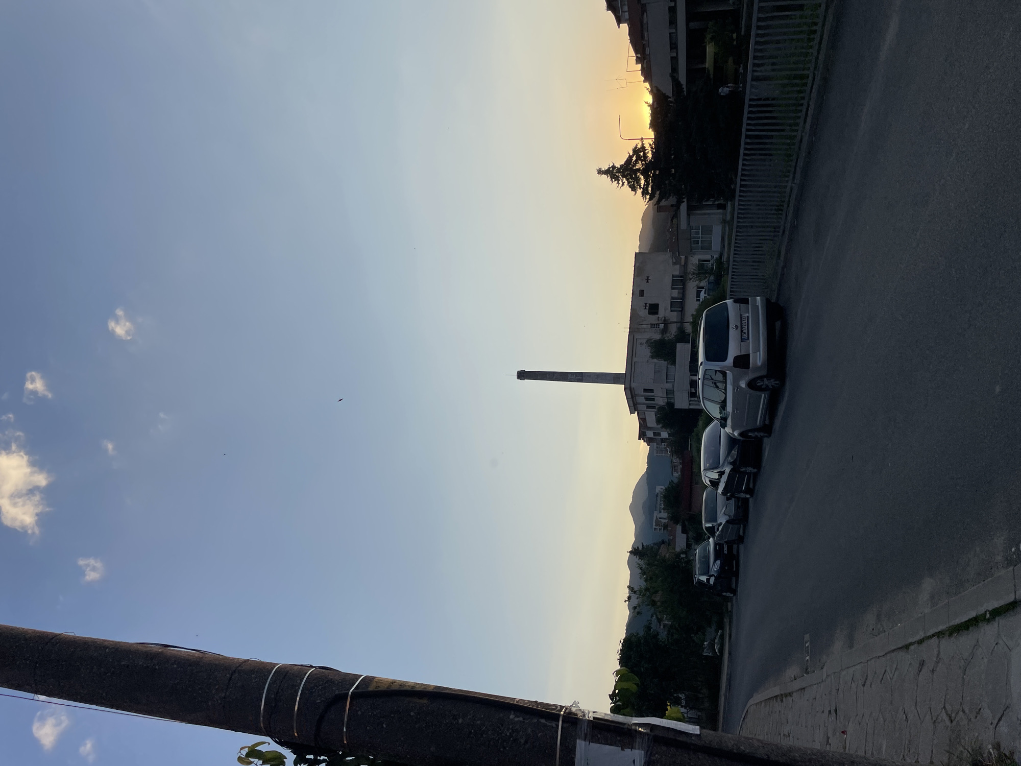 In Zlatograd, Bulgaria, a photograph captures a street scene during sunset. A large, dark utility pole stands prominently on the left side of the frame, partially obscuring the view of the sky and distant mountains. The sky transitions from a clear blue at the top to a warm, soft yellow near the horizon, indicative of the setting sun. Several cars are parked along the side of an asphalt road, which runs from the foreground towards the middle ground. A low metal fence borders the right side of the road. In the background, various buildings are visible, including a tall industrial chimney. The overall atmosphere is calm and serene, characteristic of the end of the day.