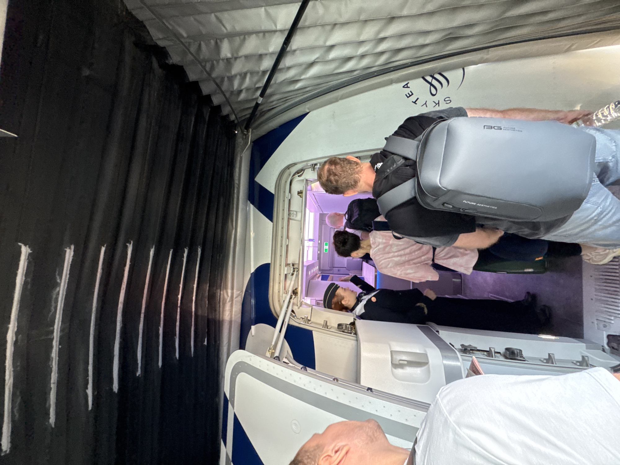 The image shows a group of people boarding an aircraft. A flight attendant in uniform stands at the door. One man has a grey backpack on. The brand of the aircraft is SKYTEA. The city and location are unknown.