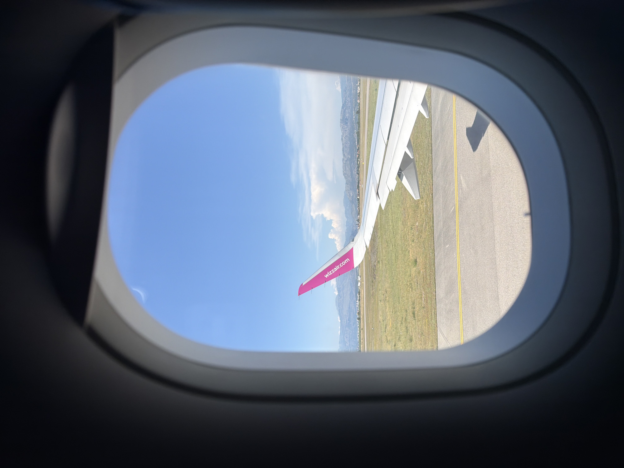 The image shows an airplane window view of an airport runway under a clear blue sky with scattered clouds. The right side of the image features a portion of the airplane's wing, which is white with a bright pink wingtip adorned with the "wizzair.com" logo. Beyond the runway, there is green grass and a distant landscape of mountains and some buildings. The perspective suggests the airplane is either on the ground at the airport or has just taken off/is about to land. The location is specified as Donja Cijevna, Montenegro.