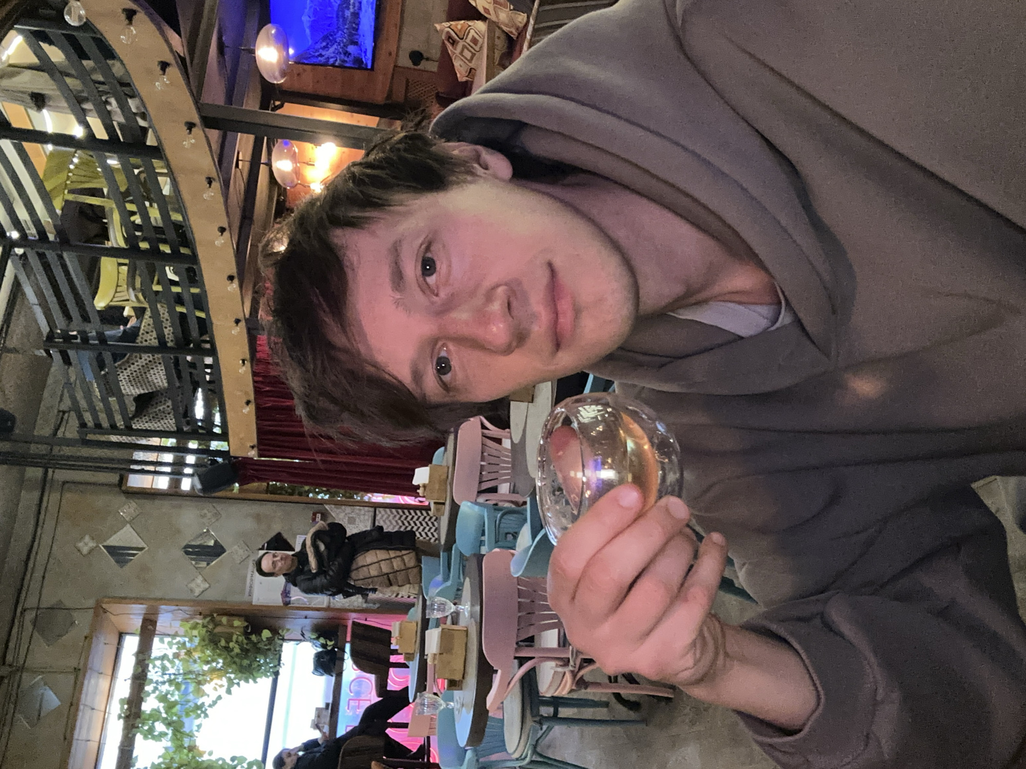 The image depicts a man in a grey hoodie, holding a glass, possibly a wine glass. He is in what appears to be a cafe or restaurant. Background details include pink and blue chairs and tables, patrons, and interior decor features. The cafe is located in Saint Petersburg, Russia.