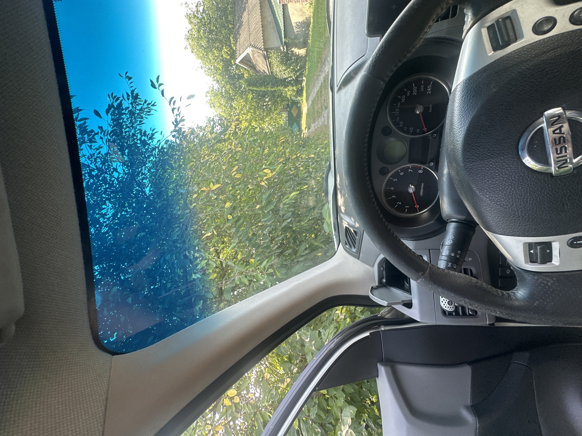 This image shows the interior of a Nissan vehicle, looking out through the windshield. The dashboard and steering wheel are visible, with the Nissan logo prominently displayed on the steering wheel. Outside the car, a dirt road leads through a rural area with trees and a house with a tiled roof. The scene is set in Shatava, Ukraine. The lighting suggests it is daytime, possibly late afternoon, given the warm sunlight filtering through the trees.