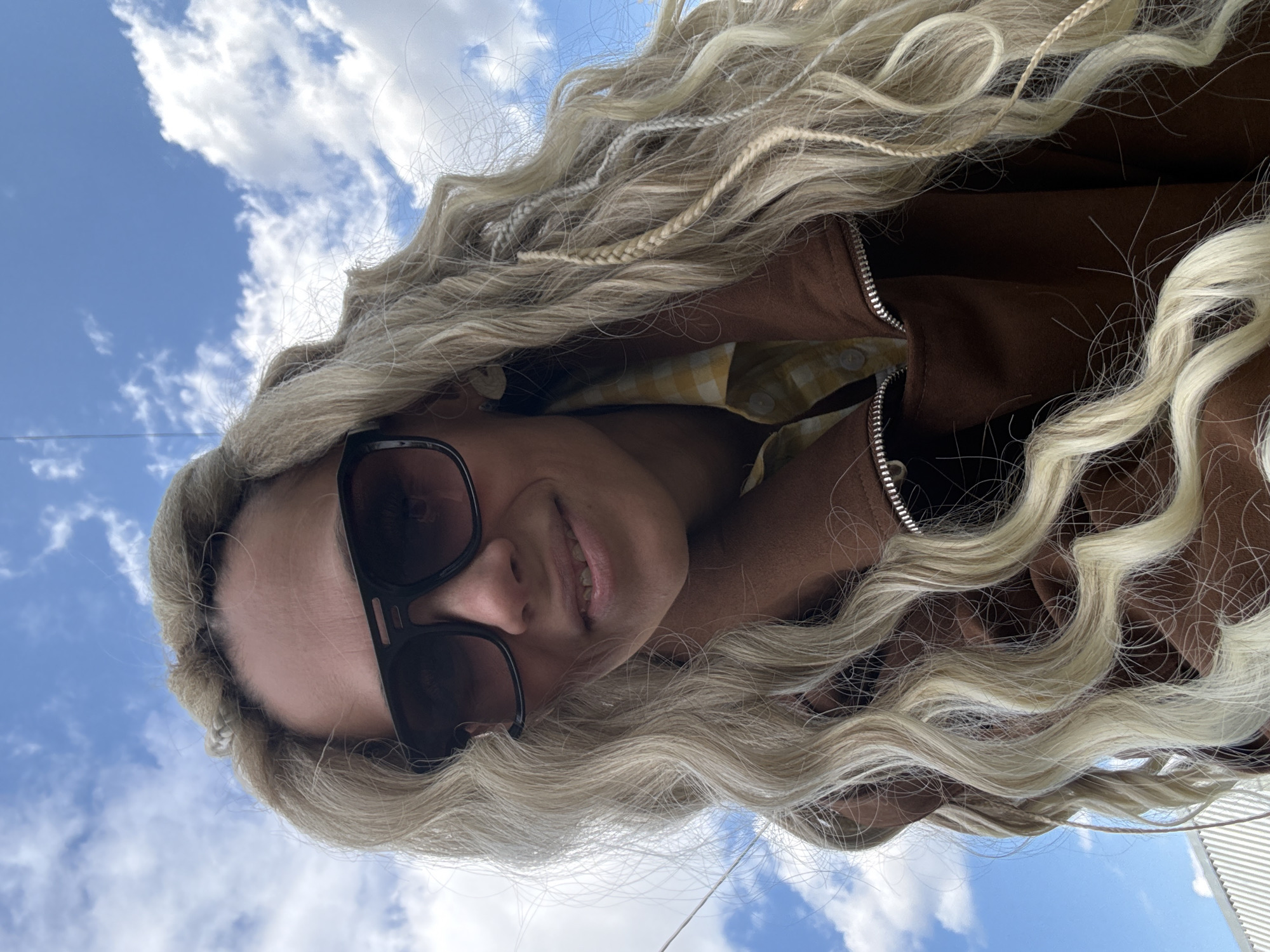 An upward-angled selfie captures a person with very long, wavy, light blonde hair, some of which is styled into thin braids. The individual is wearing dark sunglasses, which obscure their eyes but reveal a subtle smile. They are dressed in a brown jacket with a visible silver zipper, over a yellow and white plaid shirt. The original image is rotated, showing the person's head tilted towards the left, with their face partially framed by their hair.
The background is a clear blue sky filled with white, fluffy cumulus clouds, indicating an outdoor setting during the daytime. The lighting suggests a sunny day with good visibility. The angle of the shot, looking up at the sky, creates a sense of openness. On the far right edge of the image, a light-colored structure, possibly a building, is faintly visible, though not distinctive enough to pinpoint a precise location. The person appears relaxed and content. There is no visible text in the image. The photo was taken in Rusanovo, Russia.