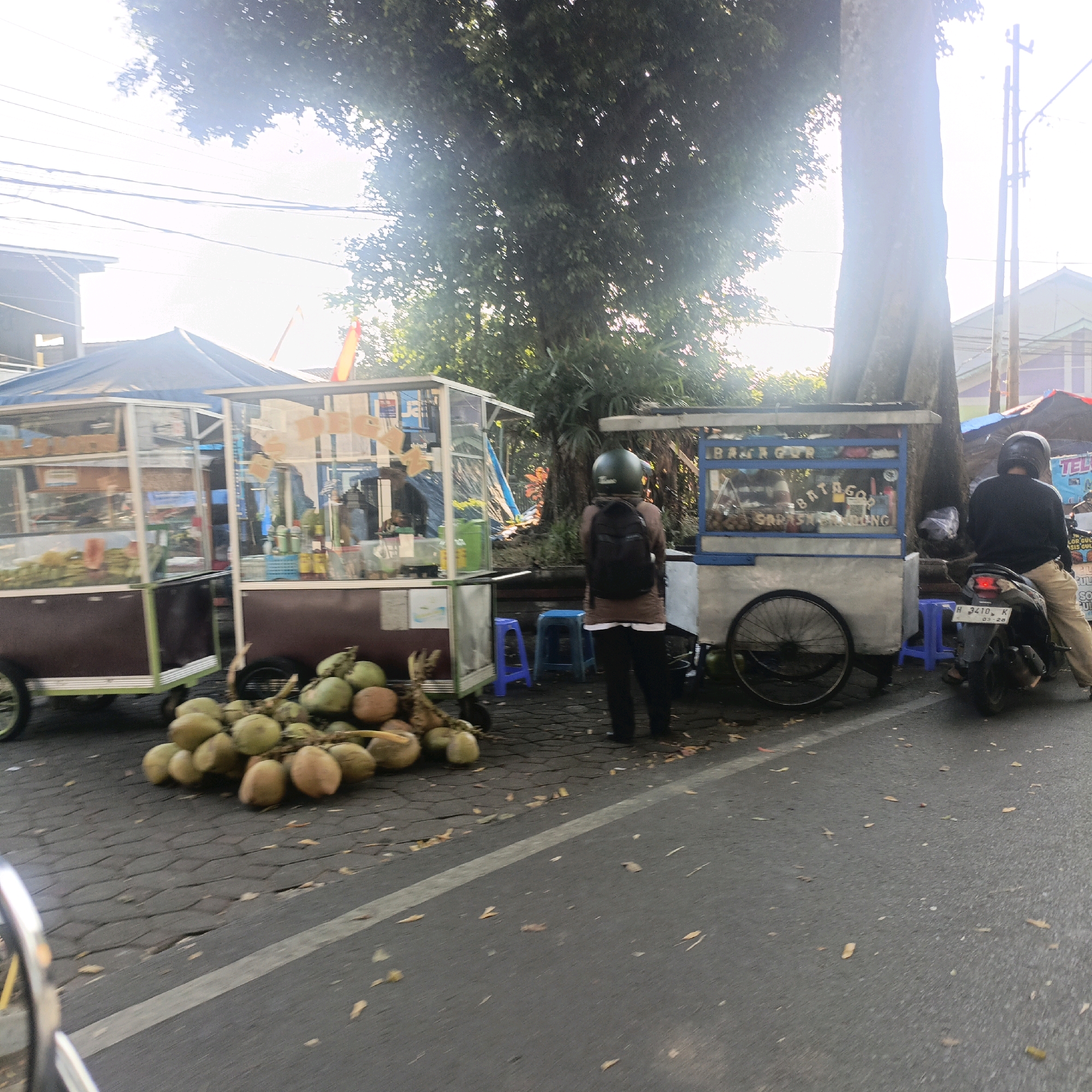 The image depicts an outdoor street scene in Salatiga, Indonesia, likely in the morning or afternoon given the bright sunlight and shadows. The foreground is dominated by a pile of coconuts on a paved surface that appears to be a street or sidewalk. To the left, a food cart with a blue awning and glass display case is visible, with various items, possibly food and drinks, inside. Further to the right, another food cart, this one with a blue frame and glass display, is positioned. A person wearing a dark helmet and a brown long-sleeved shirt and dark pants stands facing away from the camera, looking towards the food carts. Behind them, a motorcycle is parked, with another person wearing a helmet and a black shirt and khaki pants sitting on it. The background features a large tree with dense foliage, and some buildings and power lines are visible in the distance. The overall impression is that of a street-side food vending area.