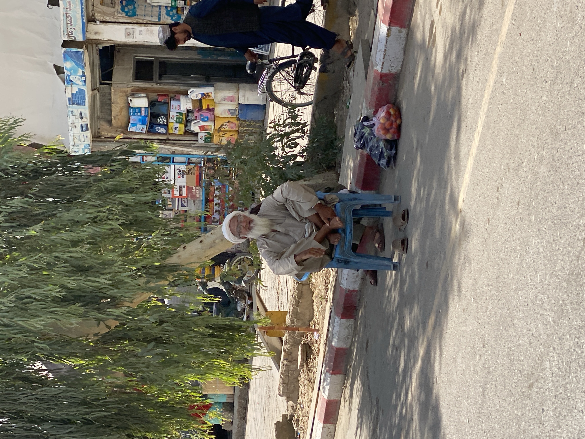 A man with a white turban and beard sits on a blue plastic chair in the shade of a tree. He wears a light-colored tunic and trousers, and sandals. In front of him, to the right, is a pile of tomatoes in a plastic bag. To the left, partially obscured by the tree, is another plastic bag. The man is sitting on a sidewalk with a red and white striped curb. Behind him, a shop is visible with shelves of bottles and containers. A man wearing a dark blue tunic and vest walks past with a bicycle. The scene appears to be a street in Kandahar, Afghanistan.