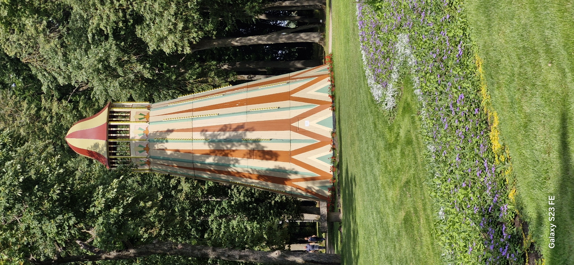 The image depicts a conical structure with a red and yellow striped roof, painted with a pattern of alternating brown and teal stripes and geometric shapes. The structure is surrounded by trees and a manicured lawn with flowerbeds. Two people are visible walking on a path in the background. The scene appears to be a park or garden. The presence of a prominent, brightly colored structure suggests it might be a recreational or historical landmark within the city of Pakruojis, Lithuania. The overall impression is of a well-maintained outdoor space during daylight hours.