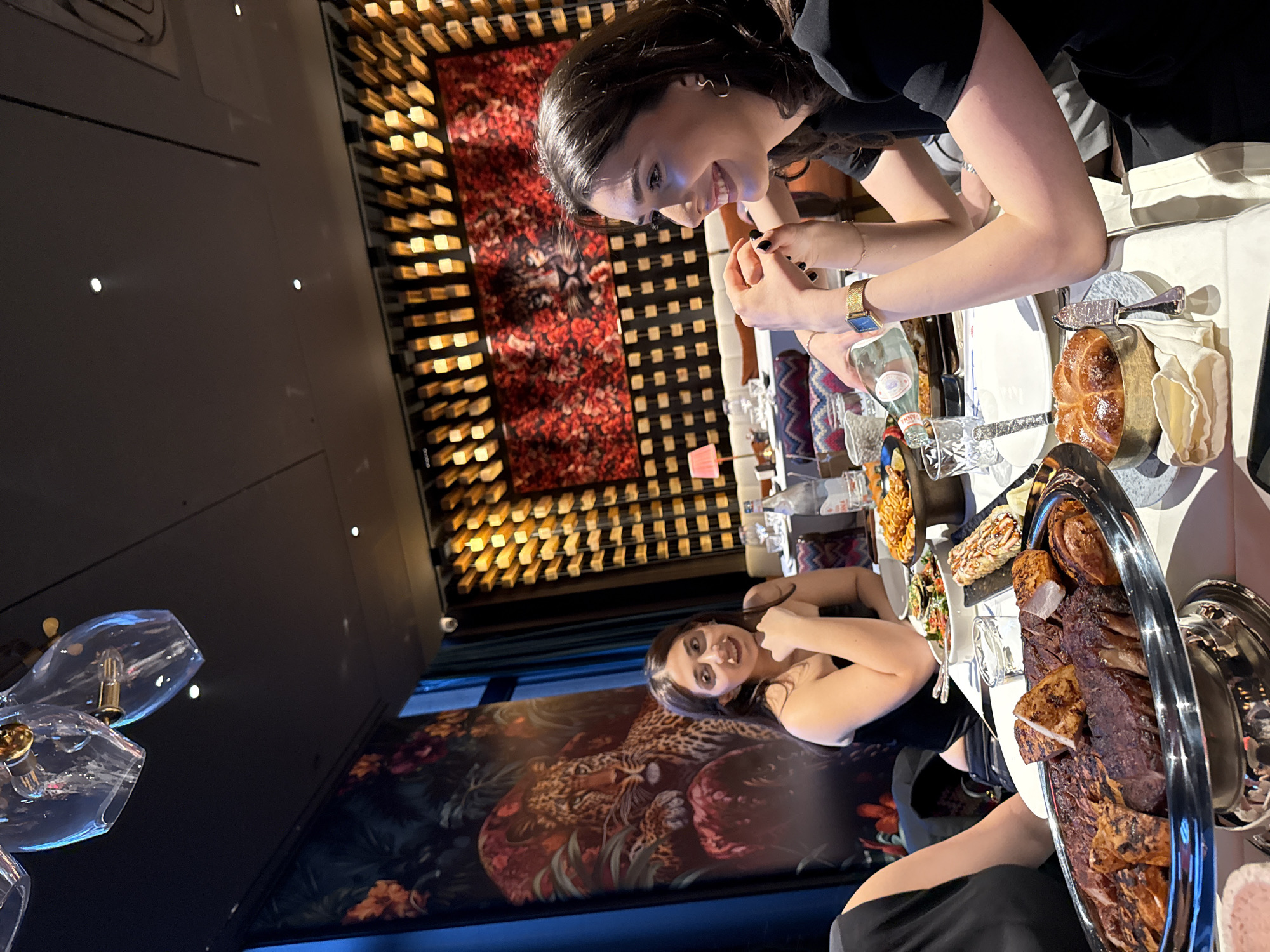 The image shows a high-angle view of a restaurant table setting with two women seated at it. The location is presumably in Yerevan, Armenia (based on the assumption from the context).
The table is covered with a white tablecloth and laden with food. On the table are items such as bread, a bottle of water, and platters of grilled meats, salads, and other dishes.
One woman is seated on the right side of the frame, facing left. She is smiling and appears to be engaged in conversation or interaction with the other person. She is wearing a black top and a gold watch.
The other woman is seated on the left side of the frame, facing the viewer. She is wearing a black dress.
The restaurant has a modern decor with unique design features. The walls are adorned with decorative panels. A chandelier is visible in the foreground.
