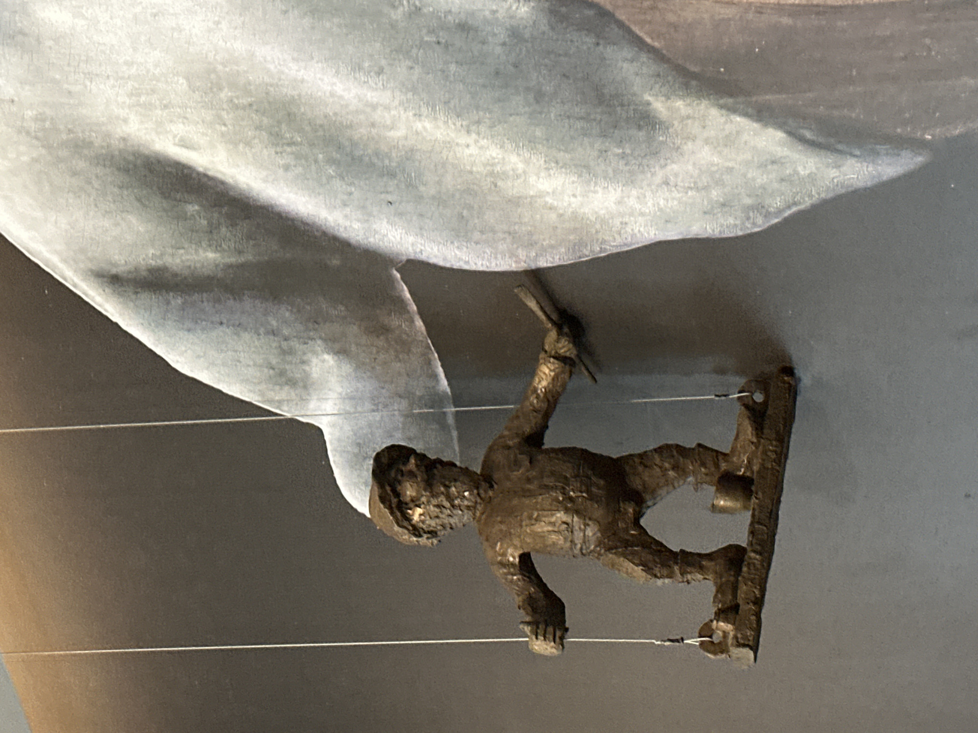 A bronze figure of a window cleaner suspended by wires is mounted on a wall. The figure is climbing on the window of a building that is the backdrop to the artwork. The figure is wearing a hardhat and holding a squeegee in its hand. It appears to be the 'Window Cleaner' sculpture located in Wrocław, Poland.