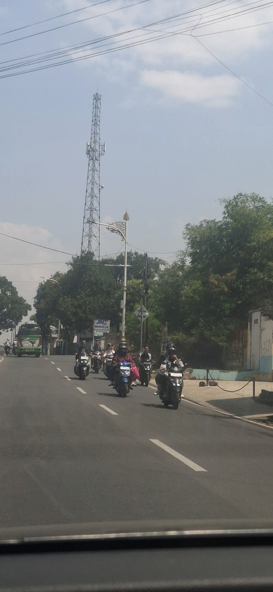On a paved road in Sukasari, Indonesia, a convoy of motorcycles with riders wearing helmets travels in the same direction. A green and white bus is visible on the left side of the road. In the background, a tall cell tower stands against a hazy sky. Trees line the right side of the road, with a wall and a building visible behind them. The scene appears to be captured from within a vehicle, with the dashboard or hood of the vehicle visible at the bottom of the frame.