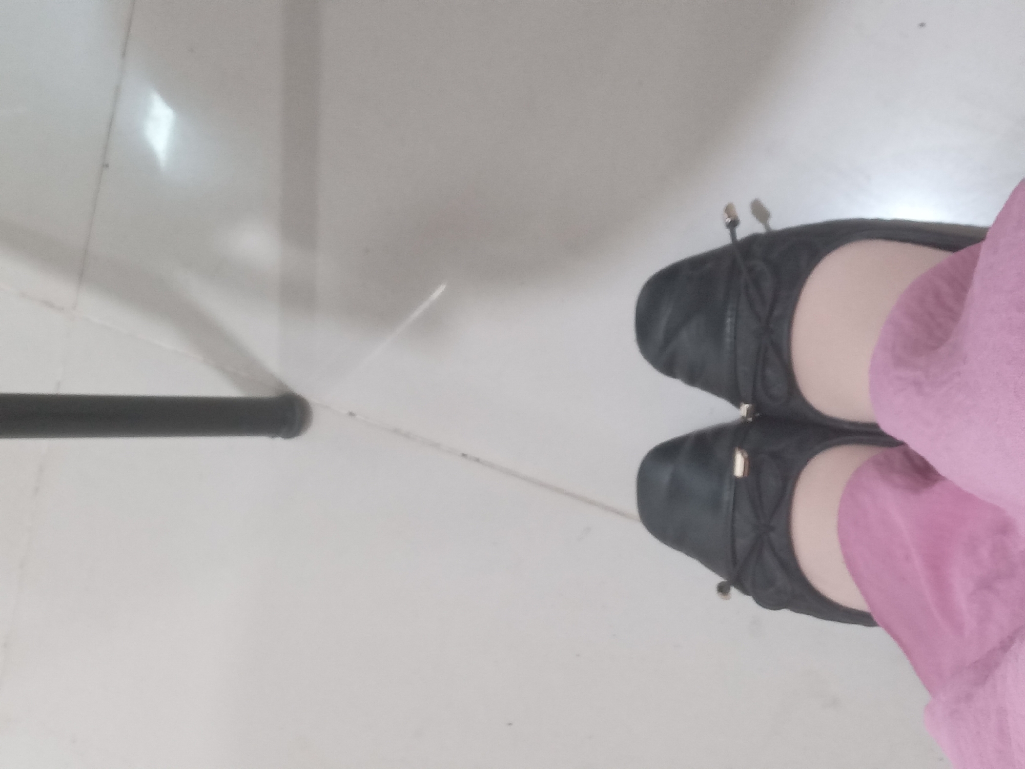The image shows a close-up view of a person's feet wearing black ballet flats and pink pants. The shoes have a decorative bow detail with gold accents. The background appears to be a tiled floor, possibly in a home or public space. The lighting suggests an indoor environment. There is a dark vertical object in the upper portion of the image, which could be a table leg or a similar furniture item. The scene is devoid of any specific urban context that would allow for pinpointing a location within Lhokseumawe, Indonesia.