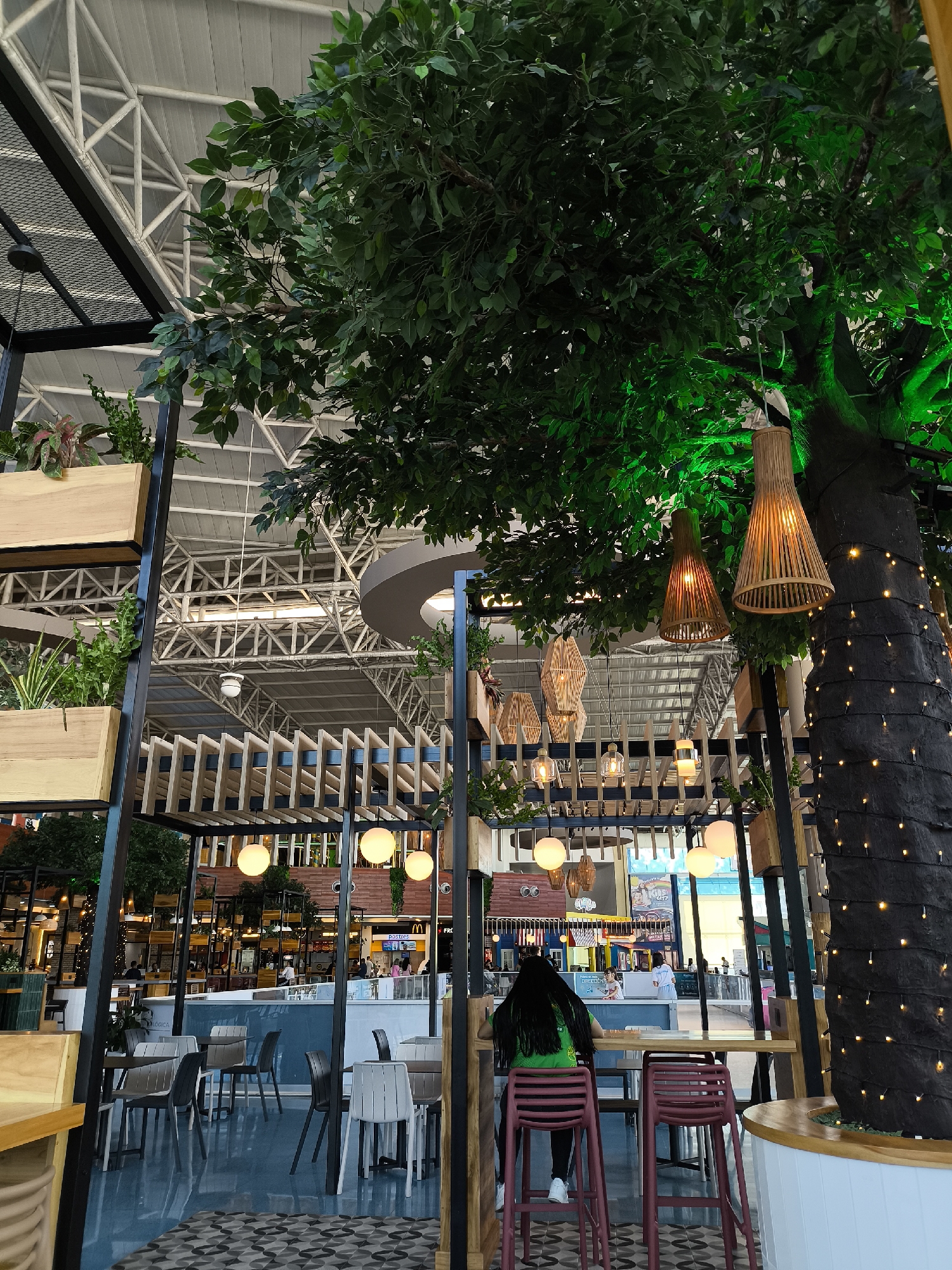 The image shows an indoor dining area, likely within a shopping center or airport. The decor features a large artificial tree with string lights and woven lampshades. Several tables and chairs are arranged throughout the space. A person with long, dark hair wearing a green shirt sits at a table. In the background, other dining areas and shops can be seen with additional people present. The location is Porlamar, Venezuela.