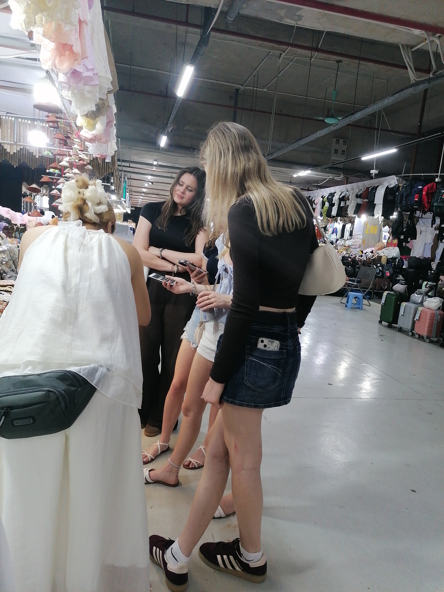 This image captures four women browsing in an indoor market, likely in Bai Chay Ward, Vietnam. In the foreground, facing away from the viewer, is a woman with intricately styled blonde hair, wearing a white, flowing halter-neck top and a dark green fanny pack. To her right, a woman with long dark hair, dressed in a black t-shirt and dark pants, looks left with a content expression, holding a smartphone. Another woman is partially visible between them, wearing a light-colored top, denim shorts, and white sandals, with an anklet on her left ankle, also holding a phone. Furthest right, seen from the back, is a woman with long blonde hair, wearing a black long-sleeved crop top, a denim mini-skirt, and a white shoulder bag. A phone with a distinct case is tucked into her back pocket. She wears white socks and black sneakers, identified as Adidas Samba by the visible "adidas" on the stripes and partially "SAMBA" text.
The market environment features a utilitarian concrete ceiling with exposed pipes, ducts, and fluorescent strip lights. Numerous pastel-colored, ruffled garments and rows of traditional conical hats hang from the ceiling, adding to the market's character. In the background, racks display various clothing items and backpacks. A blue plastic stool is on the floor, and several suitcases (green, silver, pink) are lined up, suggesting goods for sale. The floor is a light-colored, plain concrete surface. The women appear to be engaged in browsing or social interaction, with some using their mobile phones.