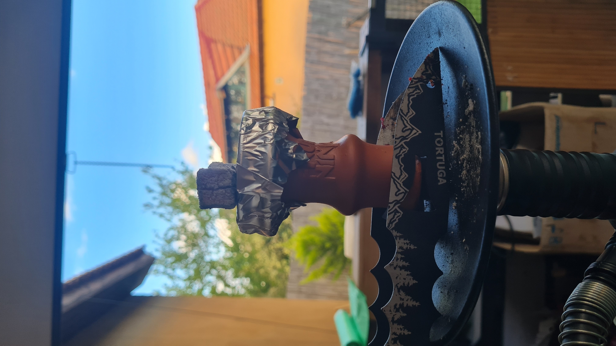 A close-up, rotated image depicts a hookah with a lit coal on top, likely set outdoors. The hookah features an orange clay bowl wrapped in foil, holding the burning coal. The stem of the hookah is black with a decorative round piece labeled "TORTUGA" adorned with a forest scene. The background shows a blurry outdoor scene with a blue sky, foliage, and buildings with red tiled roofs. The location is in Lachish Regional Council, Israel based on contextual information. The overall scene suggests someone is about to smoke or is in the process of smoking the hookah outdoors.