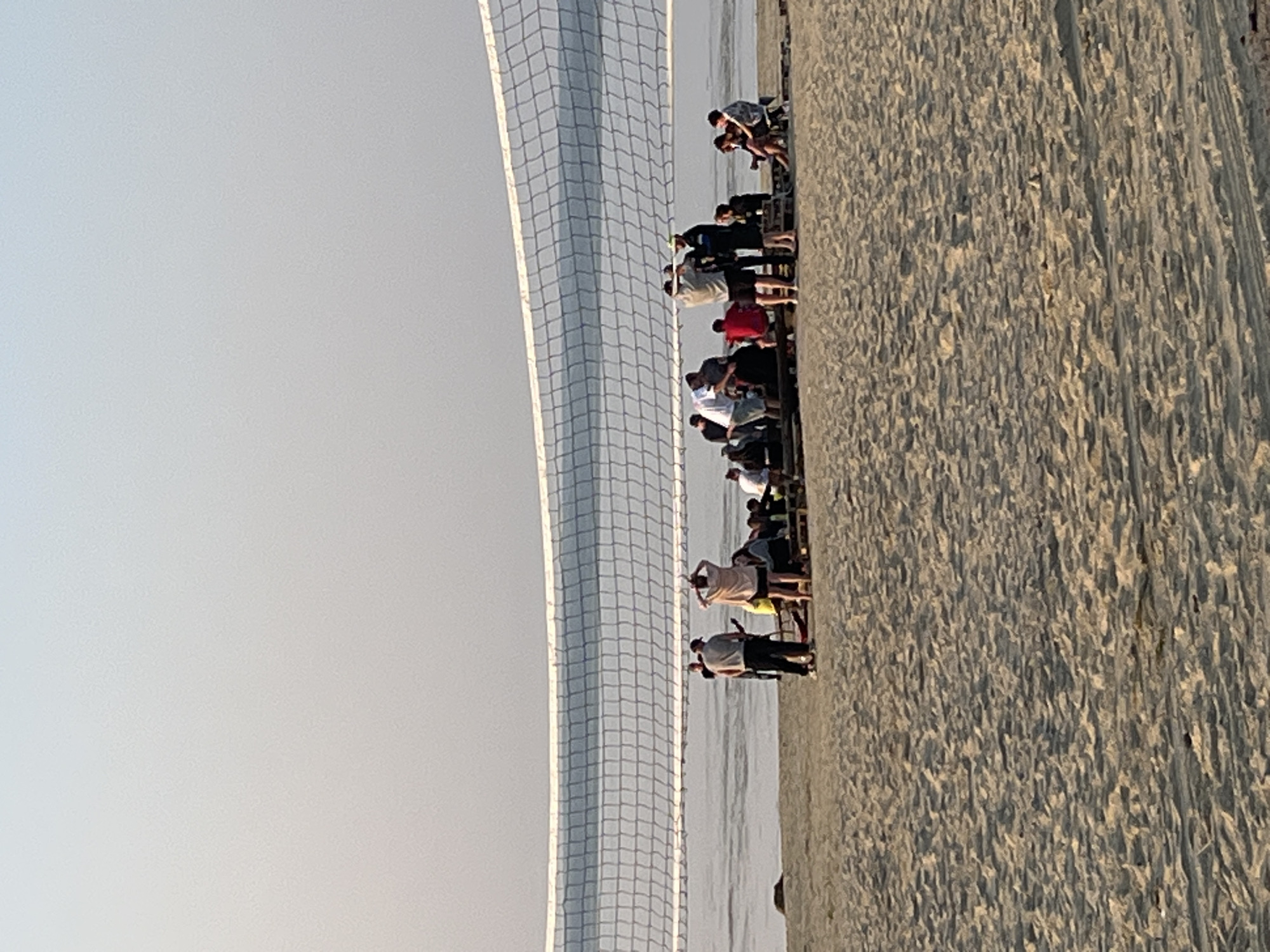A group of approximately 15 people are gathered on a sandy beach in Surakovo, Russia, near a volleyball net. The people are casually dressed and appear to be socializing or relaxing. Some are standing, while others are sitting on wooden pallets that are arranged on the sand. One person is playing a guitar. The environment includes the beach, the sea in the background, and a clear sky. The image captures a relaxed, outdoor scene.