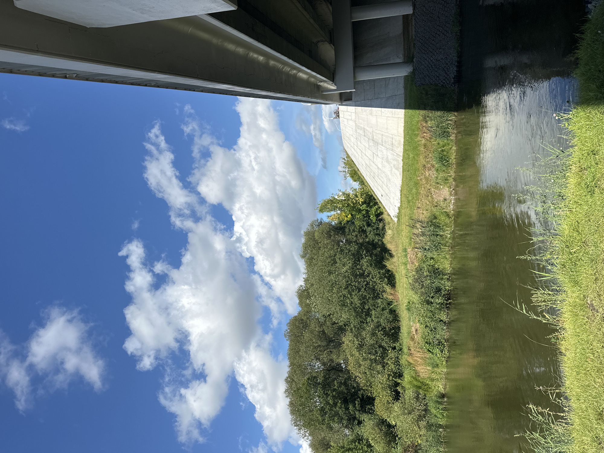 The image shows a body of water, likely a river or canal, flowing in the foreground. The water is dark green and reflects the surrounding environment. On the left bank, there is lush green vegetation, including tall trees and reeds. Beyond the vegetation, a sloped concrete structure rises, and part of a large bridge is visible overhead. The sky is a bright blue with scattered white clouds. The location is identified as Minsk, Belarus.