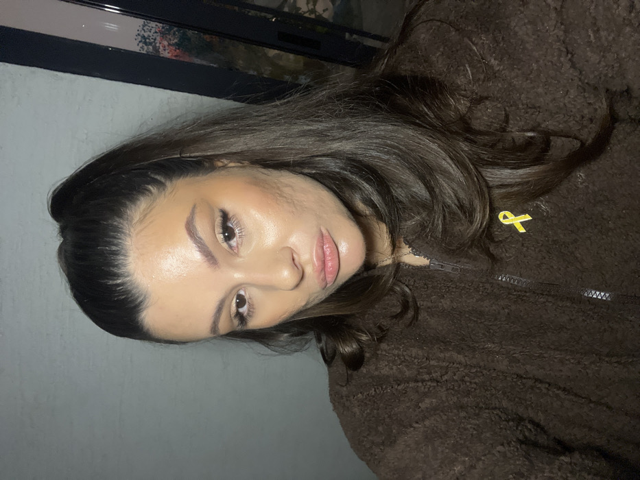 An image taken in Kiryat Bialik, Israel, depicts a female individual in a close-up, chest-up portrait. She has dark brown hair, styled in a high ponytail with loose strands cascading over her right shoulder. Her face, illuminated by frontal lighting, features prominent cheekbones, defined eyebrows, applied eye makeup, and light-colored lip gloss on slightly puckered lips. The skin on her cheeks exhibits visible texture. She wears a dark brown, textured, zip-up jacket. A bright yellow ribbon pin is affixed to the left side of her jacket, positioned approximately at chest level. The background to the viewer's left features a plain, light grey wall. To the viewer's right, a dark-framed window is visible, reflecting muted green and red hues consistent with outdoor foliage. The individual is looking directly into the camera.