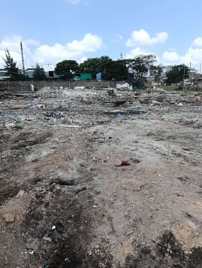 The image displays an expansive, disturbed open ground area in Nairobi, Kenya, under a blue sky with scattered white cumulus clouds. The foreground and midground are composed of uneven, dry earth, varying from light brown to grey, mixed with gravel, rocks, and a substantial amount of scattered refuse. Visible debris includes numerous plastic bags (some blue and white), fabric remnants, small fragments of wood, and general discarded materials, indicating an informal waste disposal site or neglected lot. A single reddish-brown sandal or slipper lies on the bare ground in the mid-right.
In the background, a low concrete or stone wall demarcates the rear of the littered area. Beyond this wall, various structures and vegetation are visible. On the far left, a tall, slender, light-colored industrial chimney or smokestack rises, adjacent to a dark green coniferous tree. Moving right, a line of dense, green deciduous trees partially obscures industrial-style buildings, what appear to be green shipping containers, and additional multi-story residential or commercial buildings further in the distance. Several utility poles or communication towers are also discernible against the sky in the background. No human figures or active engagement are visible in the scene.