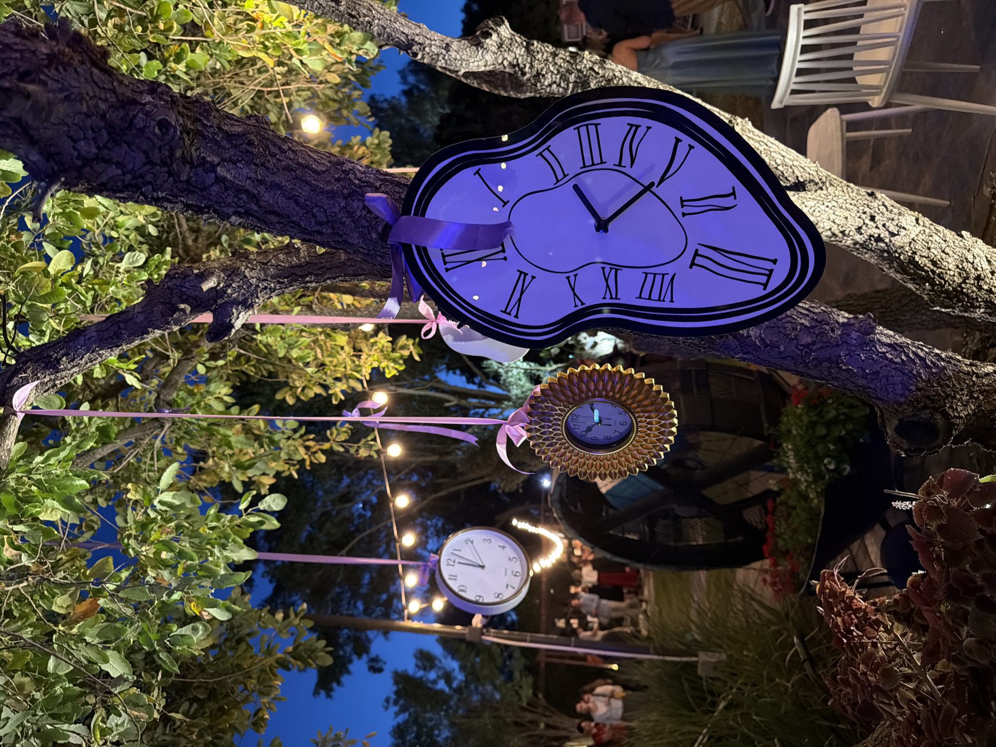 The image shows an outdoor event space at night in Nir Eliyahu, Israel. Clocks are hanging from tree branches by pink ribbons. The clocks are of various styles: a melting clock reminiscent of Salvador Dali's work, a sunburst clock, and a standard round clock. String lights are also strung through the trees. People are present in the background, standing and talking. Some white chairs are also visible.
