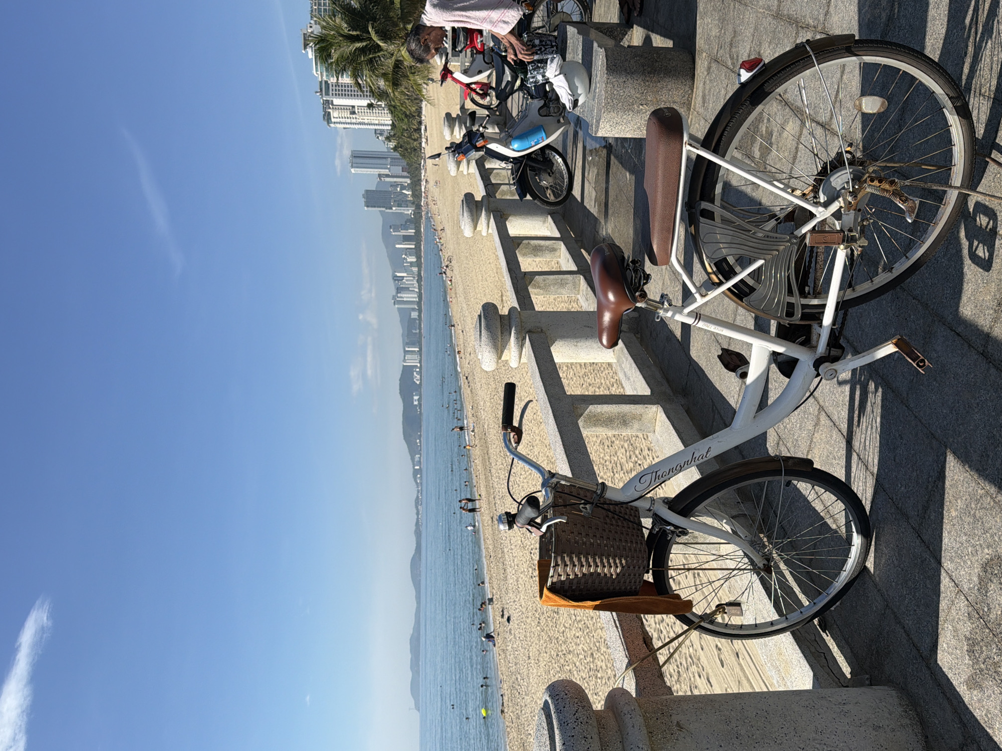 A white bicycle with a wicker basket and a brown seat is parked on a paved walkway overlooking a beach in Nha Trang, Vietnam. The bicycle is in the foreground, with its front wheel turned slightly to the left. Behind the bicycle, people are swimming and relaxing on the sandy beach, and the ocean stretches out to the horizon. In the background, a cityscape with tall buildings is visible, and palm trees line the coast. A man wearing a light-colored towel draped over his shoulders is standing near parked motorcycles. The sky is clear and blue, with a few wispy clouds. The image captures a sunny day at a popular tourist destination.