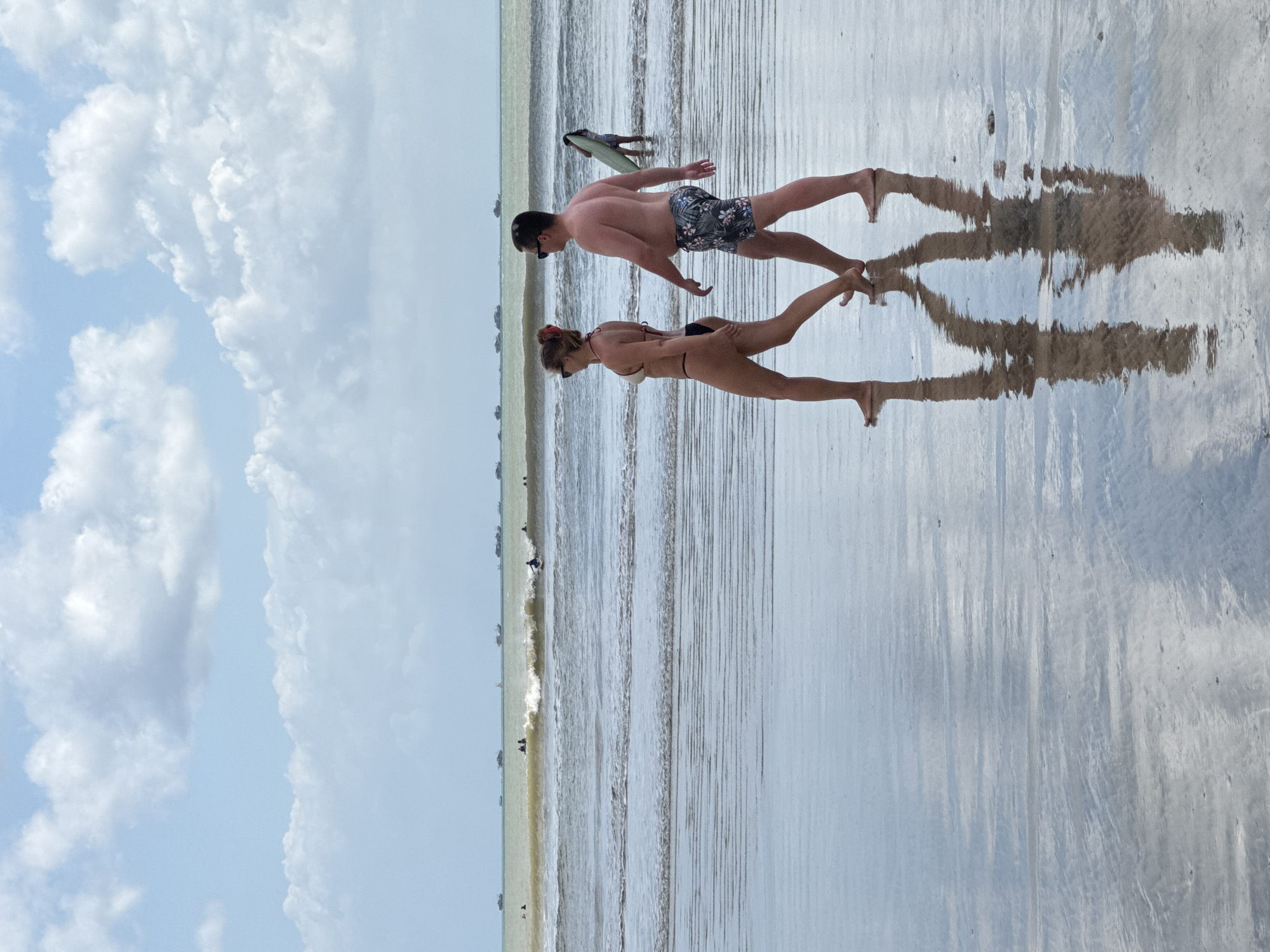 A couple walks along the wet sand of Kuta Selatan beach in Indonesia. The woman, on the left, wears a black bikini, and the man, on the right, wears patterned swim trunks. They are walking away from the camera, their reflections visible in the shallow water covering the sand. In the background, gentle waves break near a distant shoreline dotted with boats. The sky above is a light blue with scattered white clouds. Another person is visible further down the beach carrying a surfboard. The overall scene depicts a leisurely day at the beach under a cloudy sky.