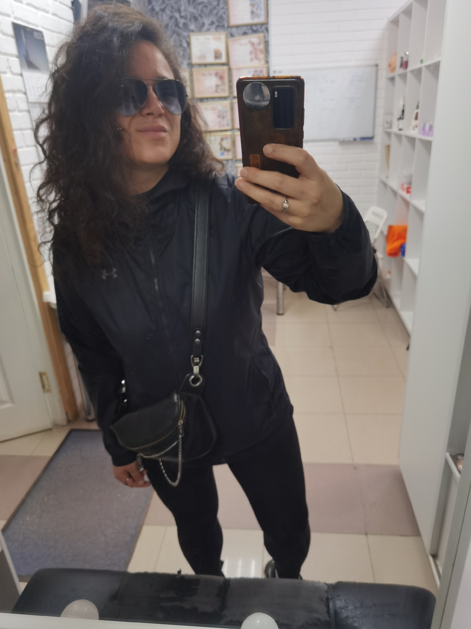 A woman is taking a mirror selfie in an indoor setting. She has dark, curly hair and is wearing aviator sunglasses, a black jacket with a small logo on the left chest, black leggings, and black athletic shoes. A black crossbody bag with a chain detail hangs from her shoulder. She is wearing a ring on her right hand.
The background appears to be a room with white brick walls and tiled flooring. On the wall behind her, there are several framed certificates or awards. To her right, there is a white shelf unit filled with various items, some of which appear to be bottles. A whiteboard is visible on the wall in the background. The lighting suggests it is daytime. The woman's expression is neutral. There is no visible text in the image. The setting appears to be a professional or retail space.