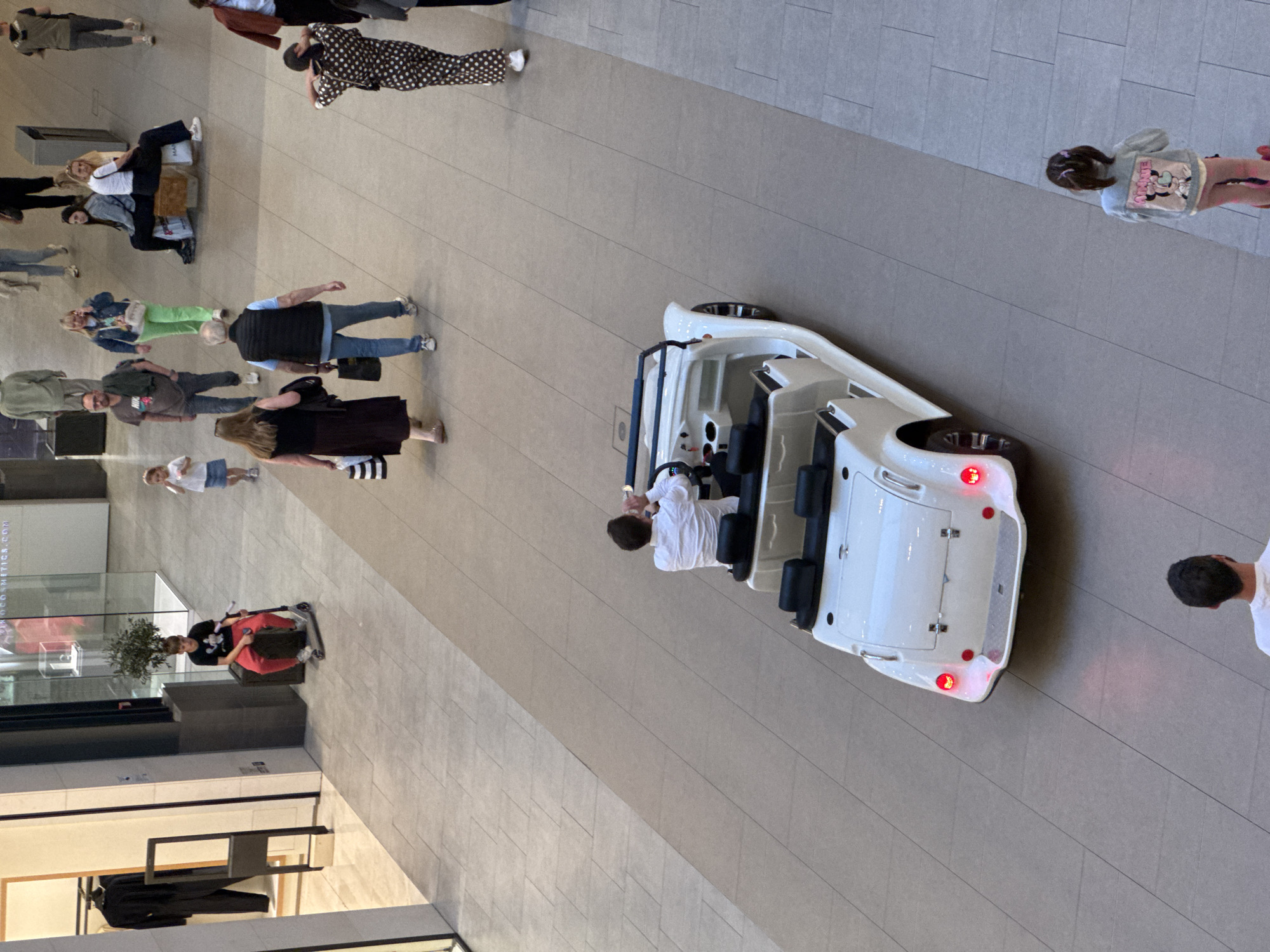 The image shows an overhead view of the interior of a shopping mall, possibly in Belgrade, Serbia. Multiple people are walking in various directions across the tiled floor. A white electric vehicle, resembling a small bus, is centrally positioned with a person in a white shirt standing beside it. A display window of a store is visible on the left side of the frame, along with clothing on display inside.