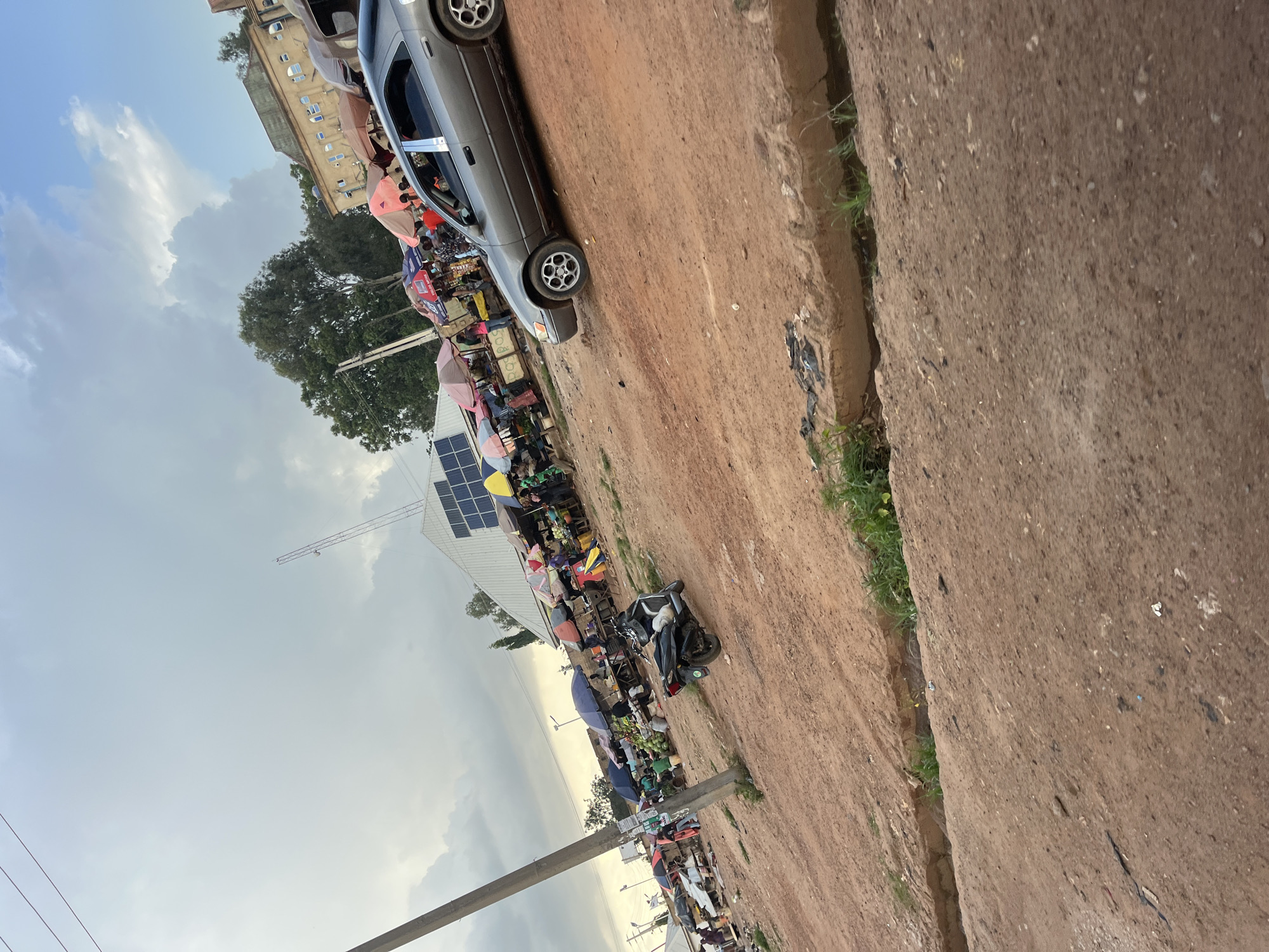 This image captures a bustling open-air market scene in Gura Top, Nigeria, likely during daylight hours under a cloudy sky. A dusty, reddish-brown dirt road dominates the foreground, with a shallow, overgrown ditch running along the right side. In the mid-ground, the market is characterized by numerous stalls covered by an assortment of colorful umbrellas, shading goods and people. Several individuals are visible, engaged in shopping or selling activities.
Prominent in the scene is a silver sedan driving on the dirt road, with its tires on the right side of the vehicle visible. A black motorcycle is parked near the market stalls. Behind the market, a building with a light-colored facade and windows is visible, suggesting a permanent structure associated with the market area. A large antenna tower stands in the background. The presence of solar panels on the roof of a building in the market further indicates modern infrastructure integrated into the traditional setting. The overall impression is one of a vibrant, active marketplace within a somewhat undeveloped urban environment.