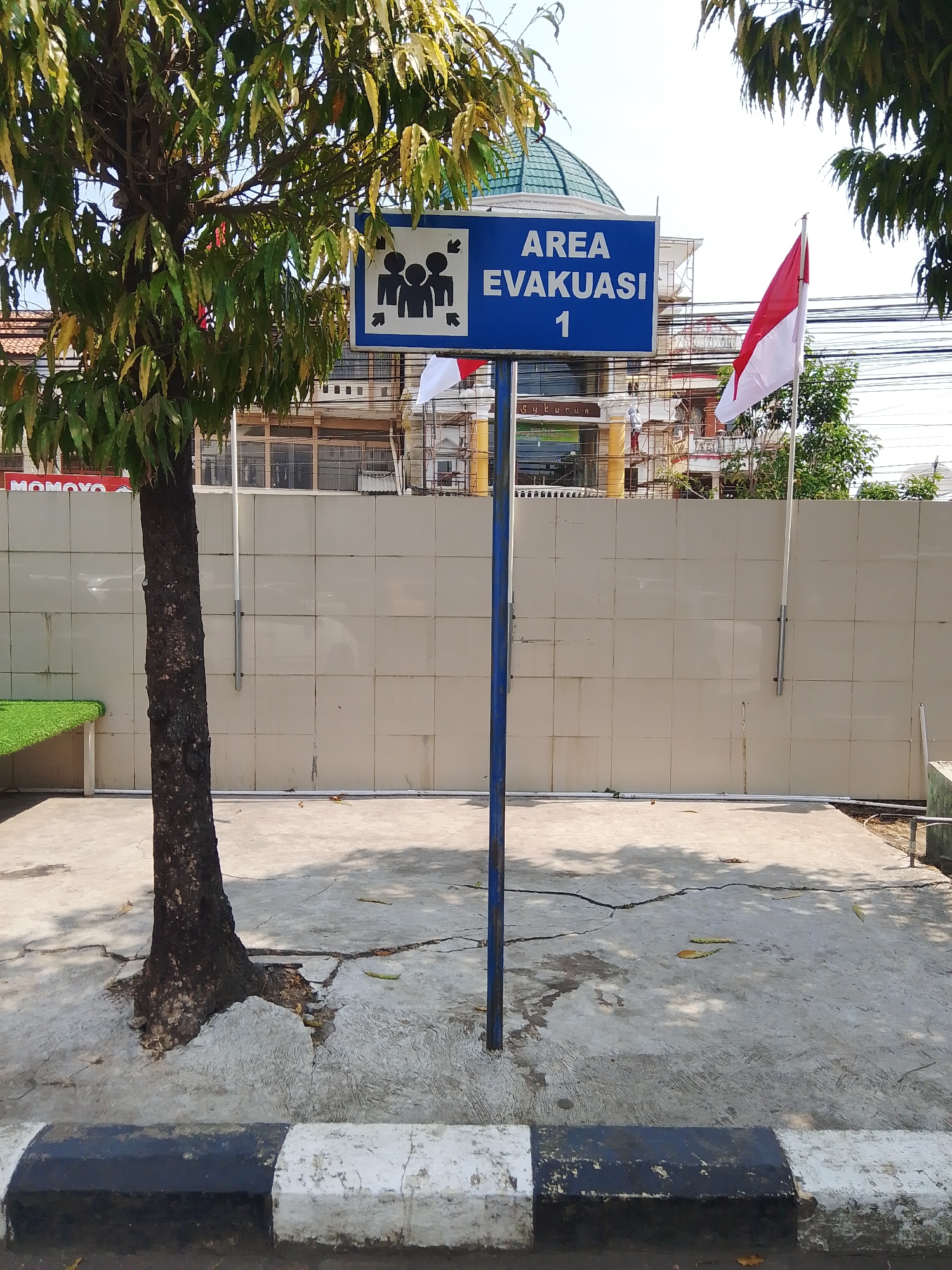 A blue sign with white lettering indicates "AREA EVAKUASI 1" and displays a pictogram of three people with arrows pointing inward. The sign is mounted on a dark blue pole. A tree with dark bark and green and yellow leaves stands to the left of the sign. Behind the tree and sign is a light beige tiled wall. In the background, a red and white Indonesian flag flies on a flagpole. The ground is concrete, cracked in several places, and partially covered with dry leaves. A black and white striped curb runs along the bottom of the image. A building with a green domed roof and scaffolding is visible behind the flag and wall. Part of a green bench or seating area is visible to the left of the tree. The scene appears to be an outdoor public space in Potroyudan, Indonesia, possibly a designated evacuation area.