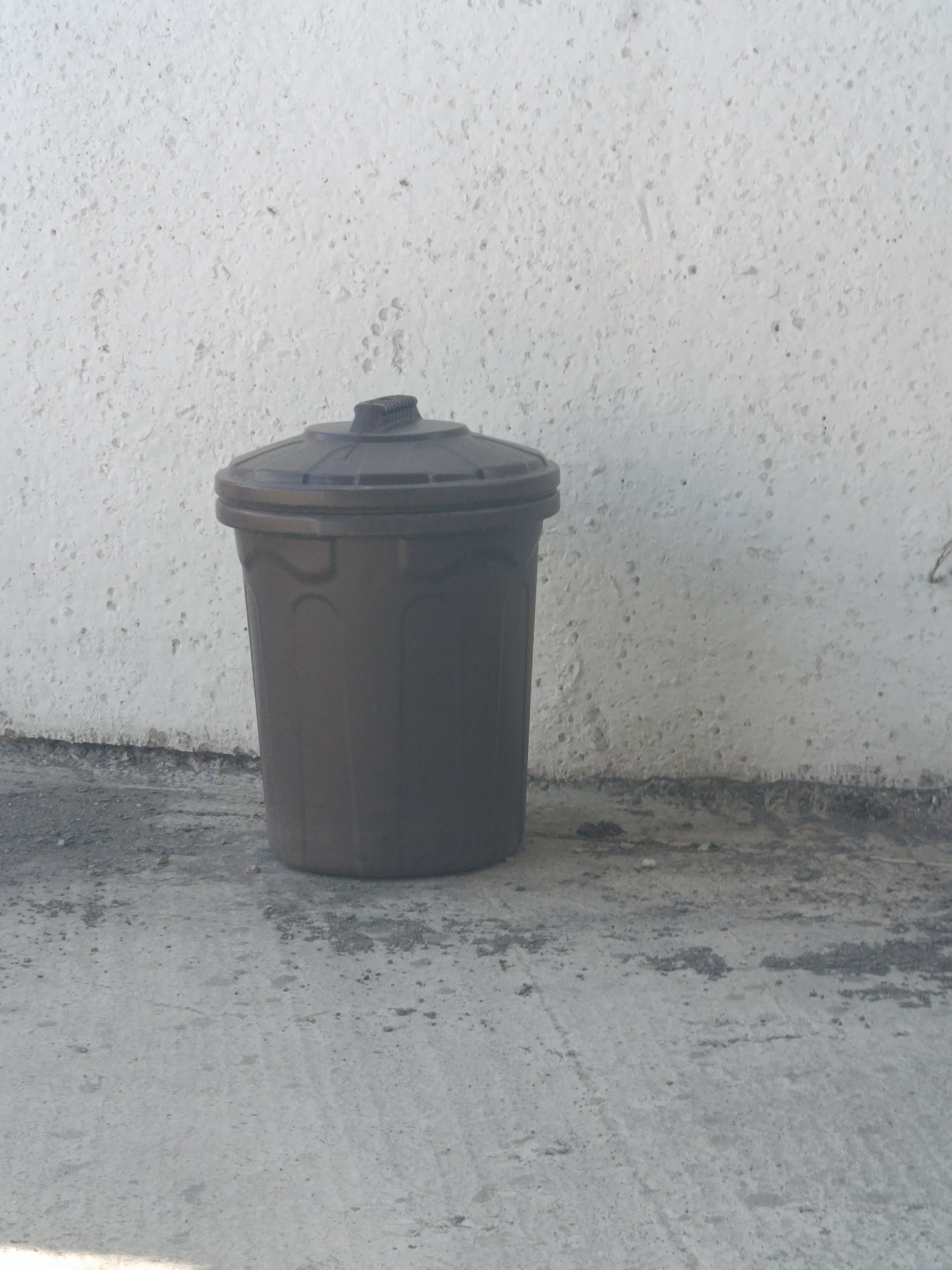 A dark brown, lidded trash can stands on a concrete surface next to a textured white wall. The trash can is cylindrical with a slightly tapered body and a domed lid that has a ribbed handle. The concrete surface is rough and uneven, with scattered pebbles and debris. The white wall behind the trash can has a coarse, speckled texture. The scene appears to be outdoors, possibly in an urban or suburban area. There are no people or animals visible in the image. The lighting suggests it is daytime. The location is identified as Mamontovo, Russia.