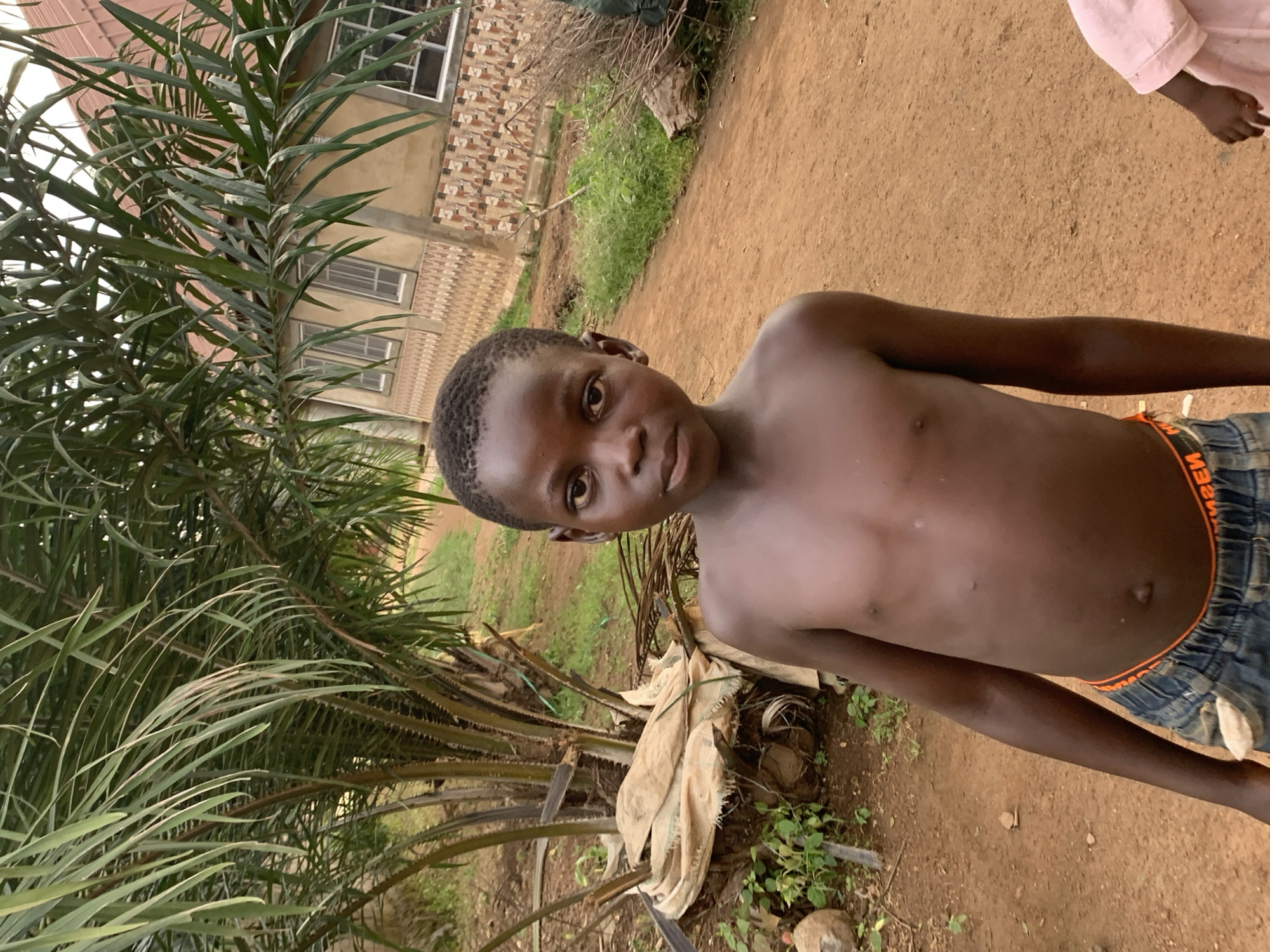 A shirtless, young Black boy with close-cropped hair stands in front of a palm tree in Agban, Nigeria. He is looking towards the camera, his body turned slightly to the left. He is wearing blue shorts with an orange waistband. Behind him are some trees and a house. The ground appears to be dirt. In the upper right corner, a small portion of a person wearing pink is visible.