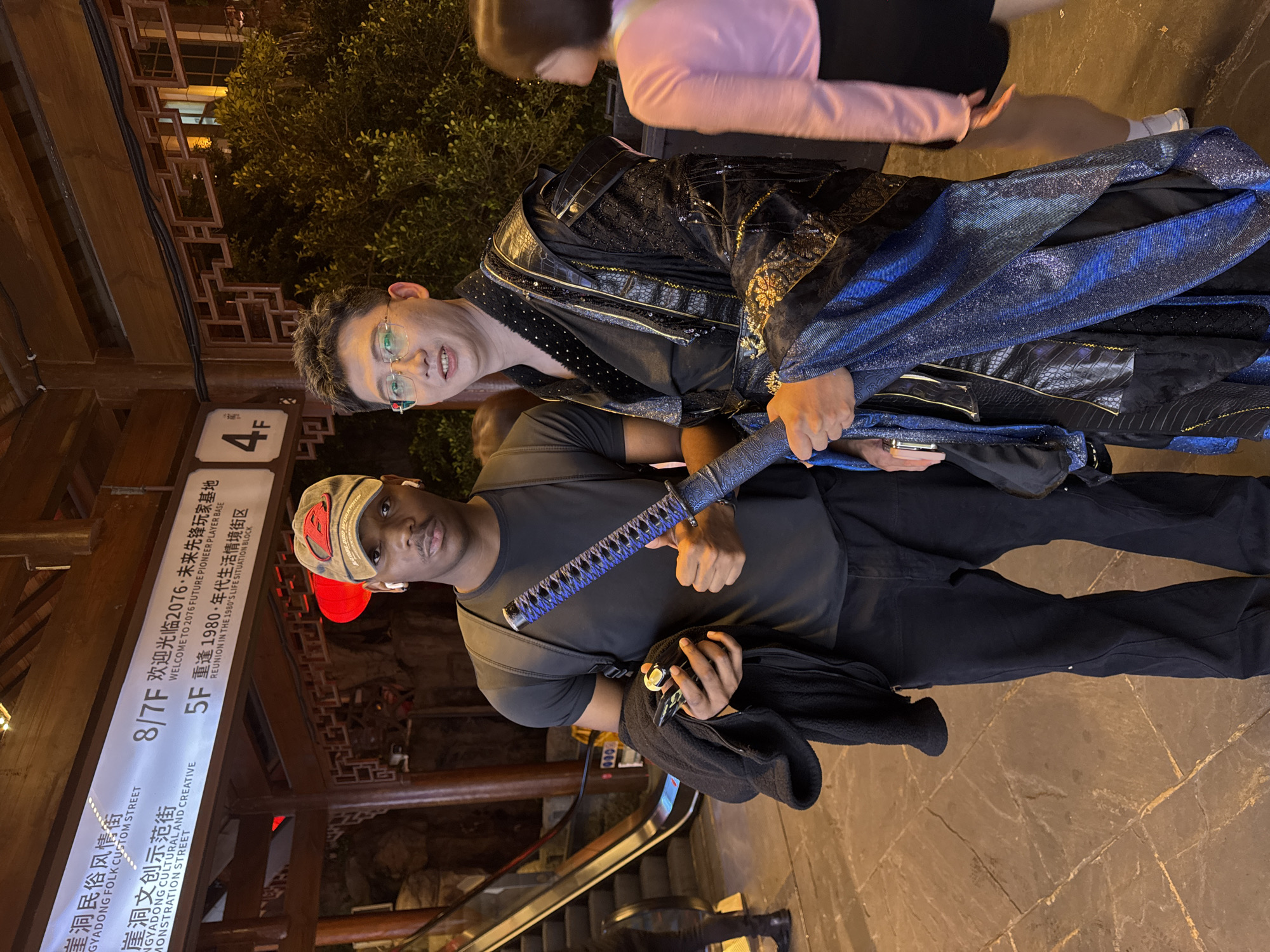 The image captures two men in Yuzhong District, China. The man on the left wears a grey t-shirt, black pants, and a cap with a red design. He holds a sheathed katana. The man on the right wears glasses and an elaborate, black and blue, possibly themed, outfit. An escalator is visible in the background. A sign displays "8/7F", "5F" and "4F" along with Chinese characters, "GYADONG FOLK CUSTOM STREET", "WELCOME TO 2076", "REUNION IN THE 1980'S LIFE SITUATION BLOCK". A third person in pink and black is partially visible in the upper right corner. The ground appears to be paved with brown stone.
