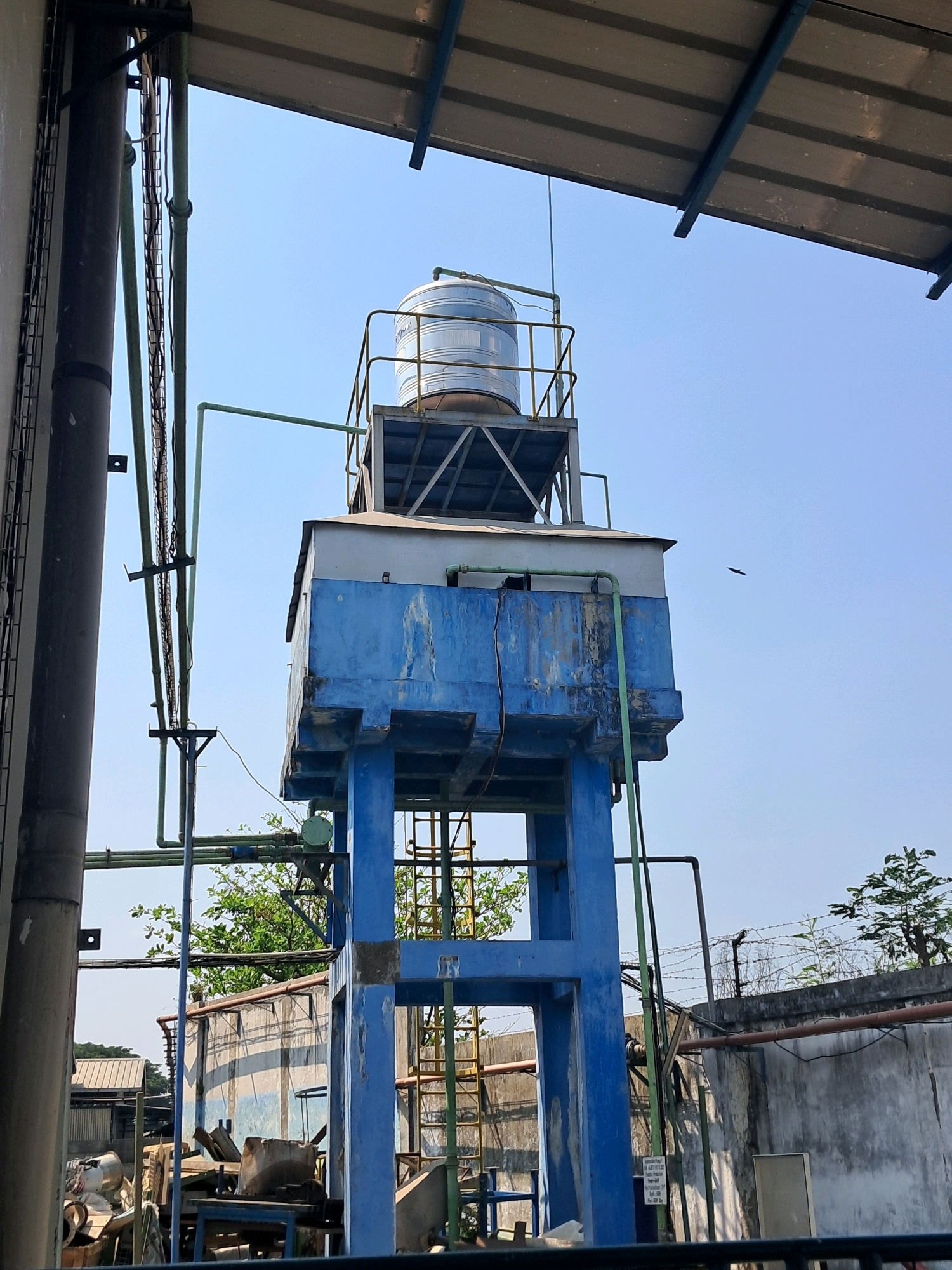An outdoor, low-angle, medium shot captures a tall, industrial water tower against a clear blue sky. The tower is a multi-level structure with a blue concrete base and legs supporting a blue, rectangular tank. A silver, cylindrical water tank sits atop this structure, surrounded by a metal railing. Green pipes run vertically and horizontally along the tower and connect to various parts of the system. To the left of the tower, a large dark pipe is visible, adjacent to a gray, textured wall. Further back, a concrete wall topped with barbed wire runs horizontally across the frame, with a tree and branches visible behind it. In the foreground, obscured by a dark railing, are some industrial materials and debris, including metal sheeting and pipes. The scene appears to be in an industrial or factory area. A small bird is visible in flight in the upper right quadrant of the sky.