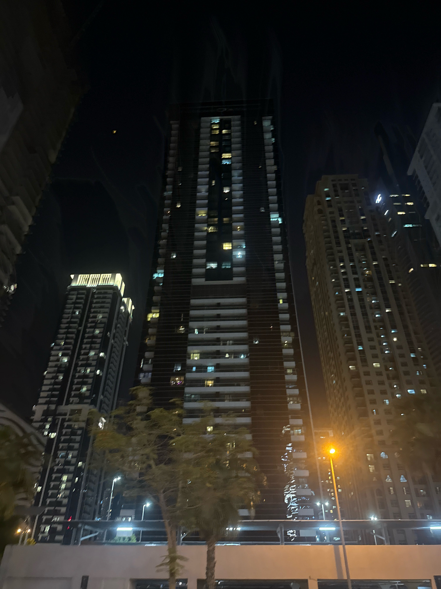 The image depicts a night scene in a modern urban environment, likely Dubai, United Arab Emirates, given the characteristic architecture. The focal point is a cluster of towering skyscrapers, illuminated by lights from their windows and external lighting. The main skyscraper in the center is tall and slender with a dark facade punctuated by many rectangular windows, some of which are lit. To its right, another skyscraper, lighter in color and broader, displays a similar pattern of illuminated windows. To the left of the central building, a third, darker skyscraper is visible, its illuminated windows creating a vertical grid. In the foreground, the tops of palm trees and other greenery are present, suggesting a landscaped urban area. A street light with a bright orange glow stands prominently to the right of center, casting light on the surrounding area. The overall impression is one of a bustling, affluent city at night, with an emphasis on verticality and artificial illumination. There are no people clearly visible in the image. The sky is dark, indicating nighttime, and there is a faint glow from the city lights reflecting upwards.