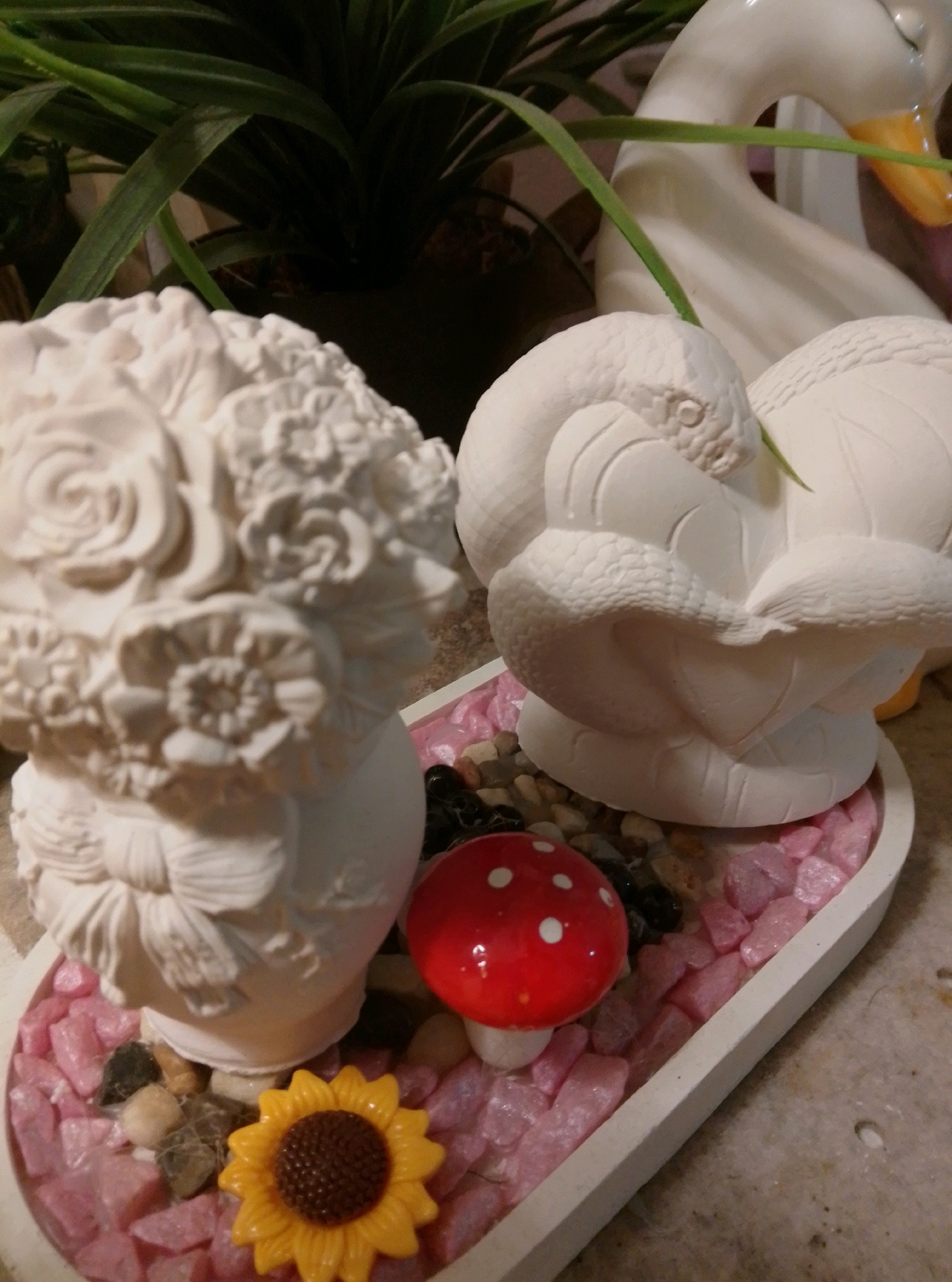 In Wedel, Germany, a white ceramic decorative piece is displayed on a textured surface. The piece is shaped like a coiled snake with its head resting on its body, and it sits within a shallow, heart-shaped white dish filled with small pink and brown stones. To the left of the snake is a white ceramic vase adorned with sculpted roses and other floral elements. In front of the vase, a small red and white polka-dotted mushroom figure is placed. To the right of the mushroom, a yellow plastic sunflower decoration is visible. In the background, a large white ceramic swan figure is present, along with green foliage from a potted plant.
