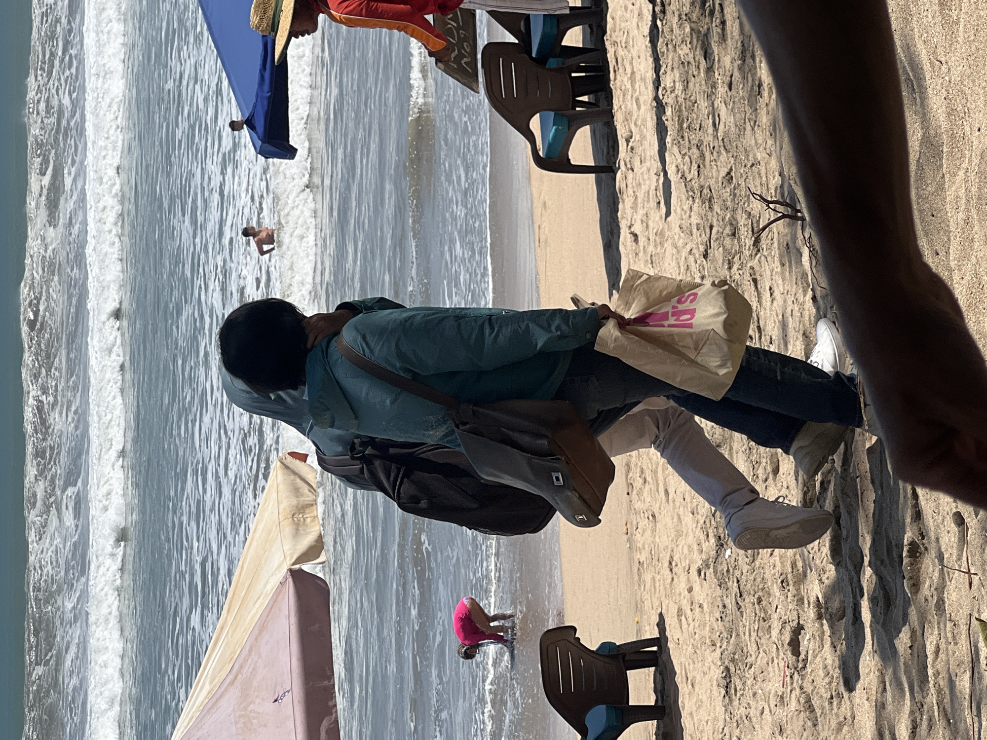 In Kuta, Indonesia, on a sunny day at the beach, two individuals are walking across the sand near the shoreline. The person in front is wearing a teal jacket and jeans, carrying a brown shoulder bag and a beige paper shopping bag with a pink logo. The person behind them is wearing lighter-colored pants and carrying a black backpack. To their left, a pink beach umbrella is set up, and to their right, a blue canopy is visible. In the water, several people are swimming and playing in the waves. A young child in a bright pink swimsuit is bent over near the water's edge, appearing to interact with the sand or water. Several lounge chairs are positioned on the beach. The ocean exhibits moderate wave activity under a bright sky.