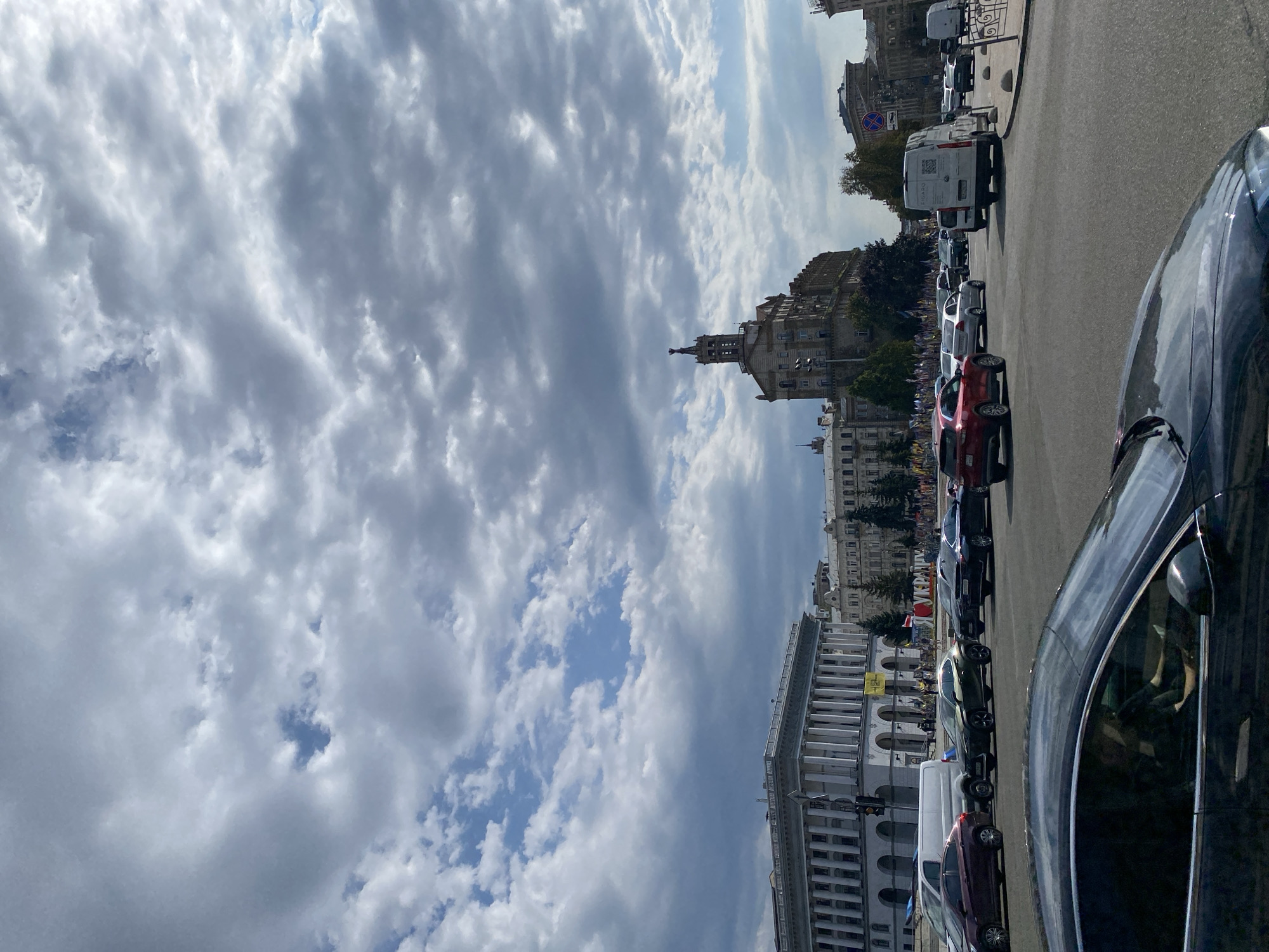 A wide-angle shot captures a bustling city scene under a dramatic, cloudy sky. In the foreground, the sleek, dark body of a car with tinted windows and a driver visible inside occupies the lower portion of the frame. Beyond this vehicle, a street is filled with a variety of cars, including a red SUV, a white van, a dark sedan, and other vehicles of different colors and makes, all navigating the intersection.
The background reveals a grand architectural landscape. On the left, a large, neoclassical building with a colonnade facade stands prominently. To its right, a distinctive, multi-story building with ornate details and a tower topped with a statue is visible. Further back, more buildings with similar architectural styles can be seen. Numerous flags, many in blue and yellow, are displayed along the street and on the buildings, suggesting a national or patriotic event. Trees are interspersed throughout the scene, adding touches of greenery. The overall impression is one of a lively urban environment with significant buildings and a notable display of flags.