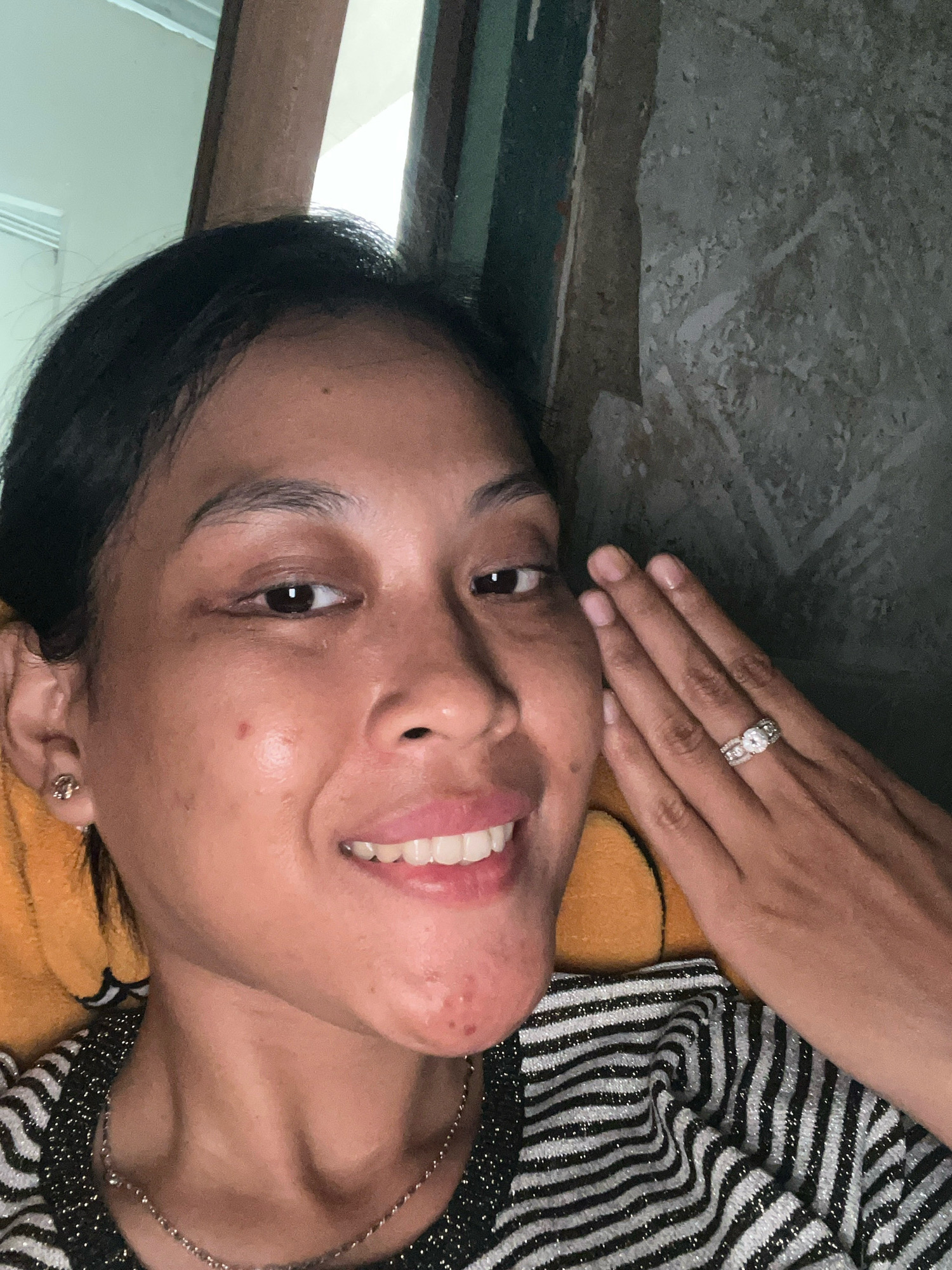 A close-up, selfie-style shot shows a woman smiling at the camera. She has dark hair pulled back, and is wearing a black and silver striped shirt. Her left hand is resting on her cheek, and her right hand is near her face, adorned with a diamond ring. The background is dimly lit and appears to be an indoor setting, possibly a room in Kecamatan Pati, Indonesia, with a textured wall and what looks like a window with light filtering through.