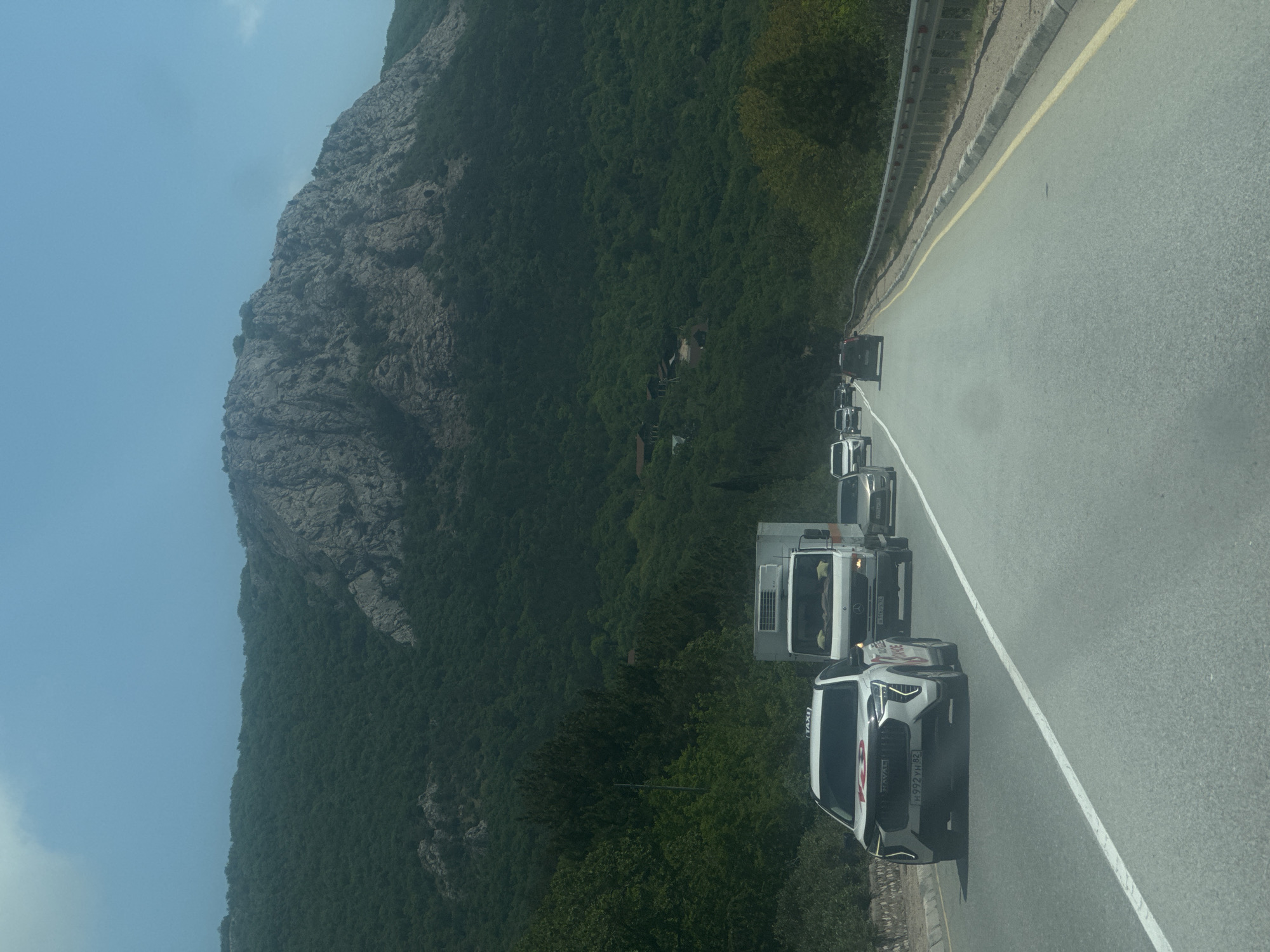 The image shows a line of vehicles on a winding road adjacent to a mountain covered in dense green vegetation. The lead vehicle is a white car with the word "TAXI" visible on its side. The other vehicles in the line include a flatbed truck and several other cars, all traveling in the same direction. The road is marked with a white line separating the lanes and a yellow line along the edge. The environment includes a large rocky mountain on one side of the road and a lush green hillside on the other. Based on the location information provided, the scene is likely in Орлиновский округ, Ukraine.
