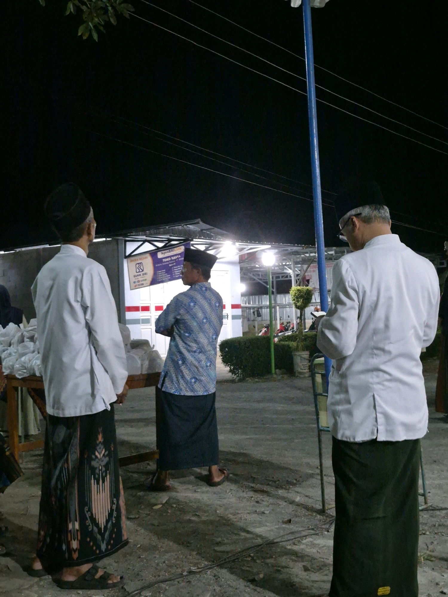 A nighttime scene in Dukuhrejo, Indonesia, captures three men standing outdoors. The man on the left, wearing a black cap and a white robe over a patterned sarong, stands with his back to the camera. Beside him, a wooden table is laden with white plastic bags. To the right, a man in a blue patterned shirt and dark sarong faces away, arms crossed. Further right, another man in a black cap and white robe, with glasses, reads from a small device. Behind them, a building with overhead lighting and a blue pole are visible, along with electrical wires and some foliage. The ground is paved with concrete and dirt.